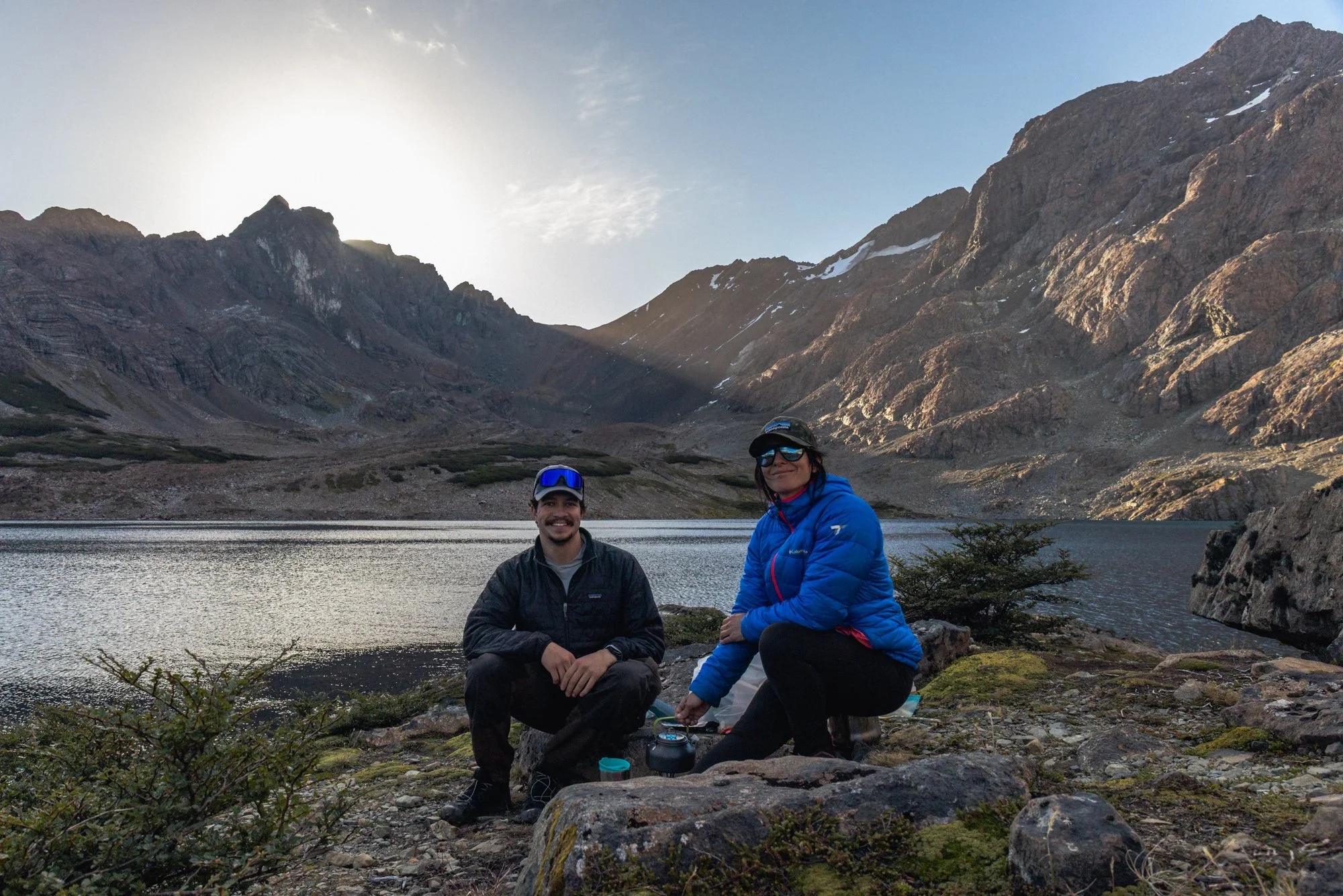 Two people sitting near a lake in a mountainous area, with rugged mountains and patches of snow in the background, during late afternoon or early evening.