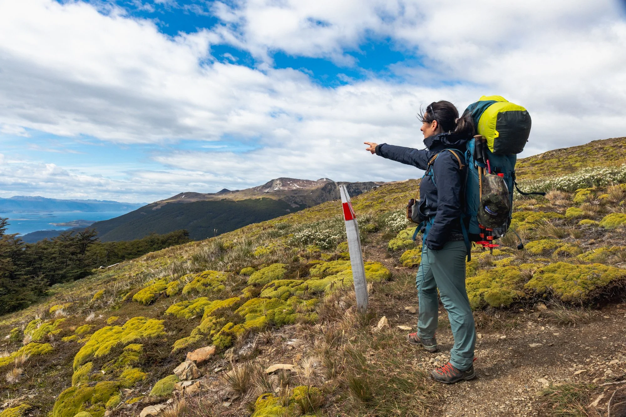 Hiker with a large backpack standing on trail pointing toward distant mountains and lake under partly cloudy sky.