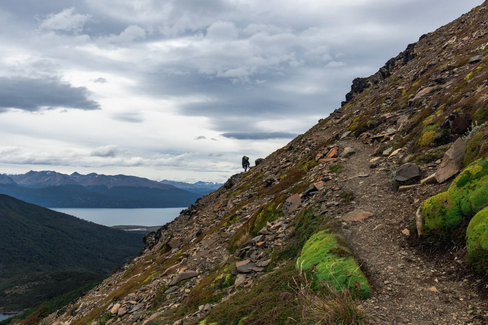 A hiker walking along a narrow trail on the side of a mountain with rocky terrain and patches of green moss, overlooking distant mountains and a body of water under a cloudy sky.