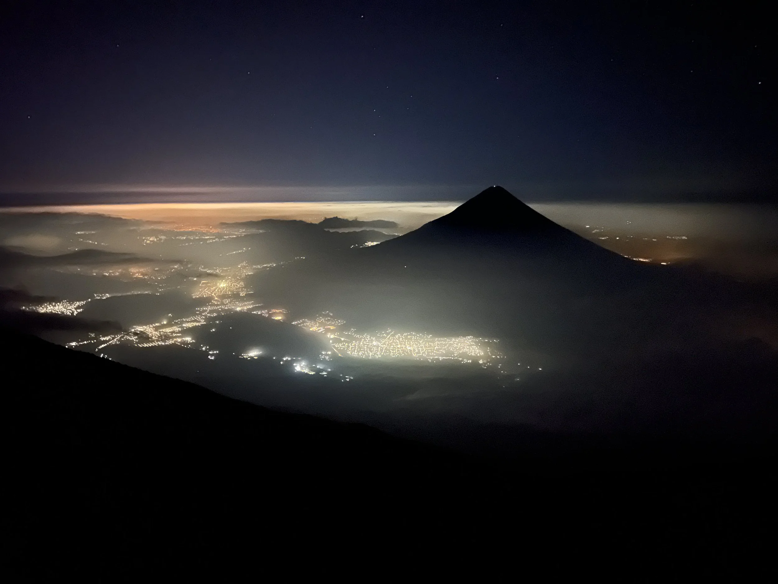 Nighttime view of a mountain with a conical shape, surrounded by city lights and fog, under a starry sky with a horizon glow.