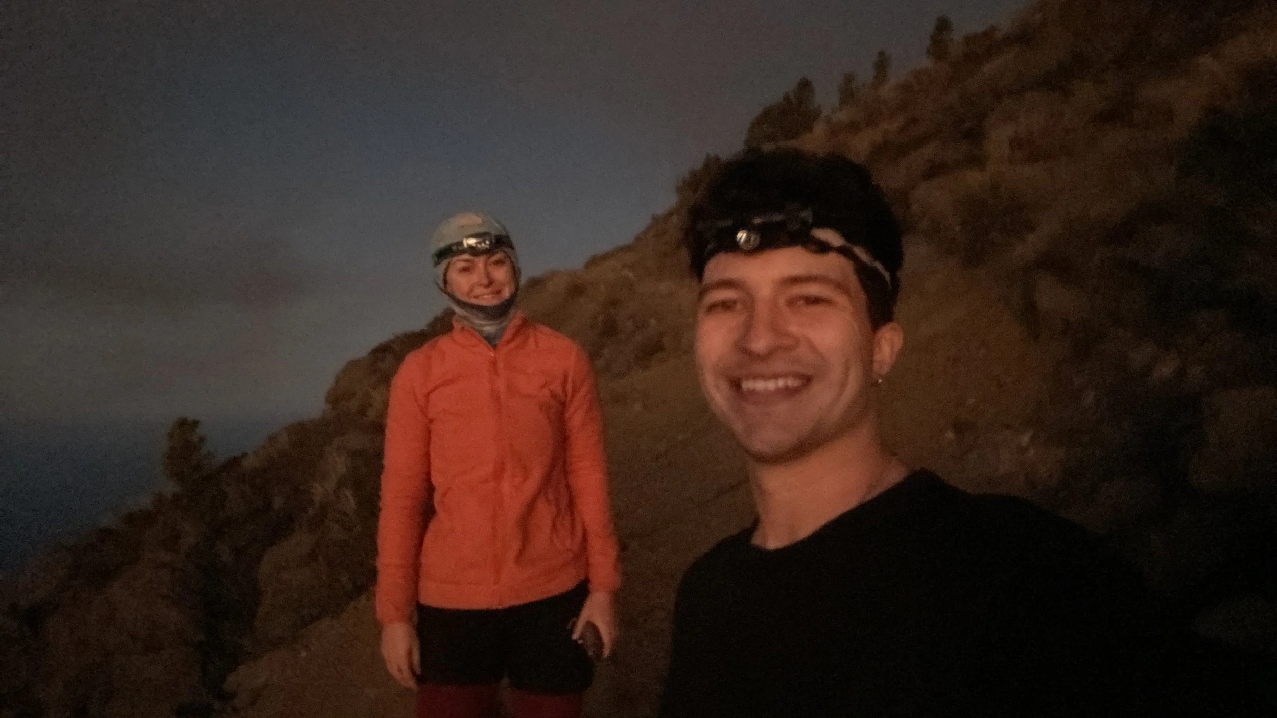 A smiling man and woman taking a selfie outdoors at dusk, wearing headlamps and outdoor gear, with a rocky hillside in the background.