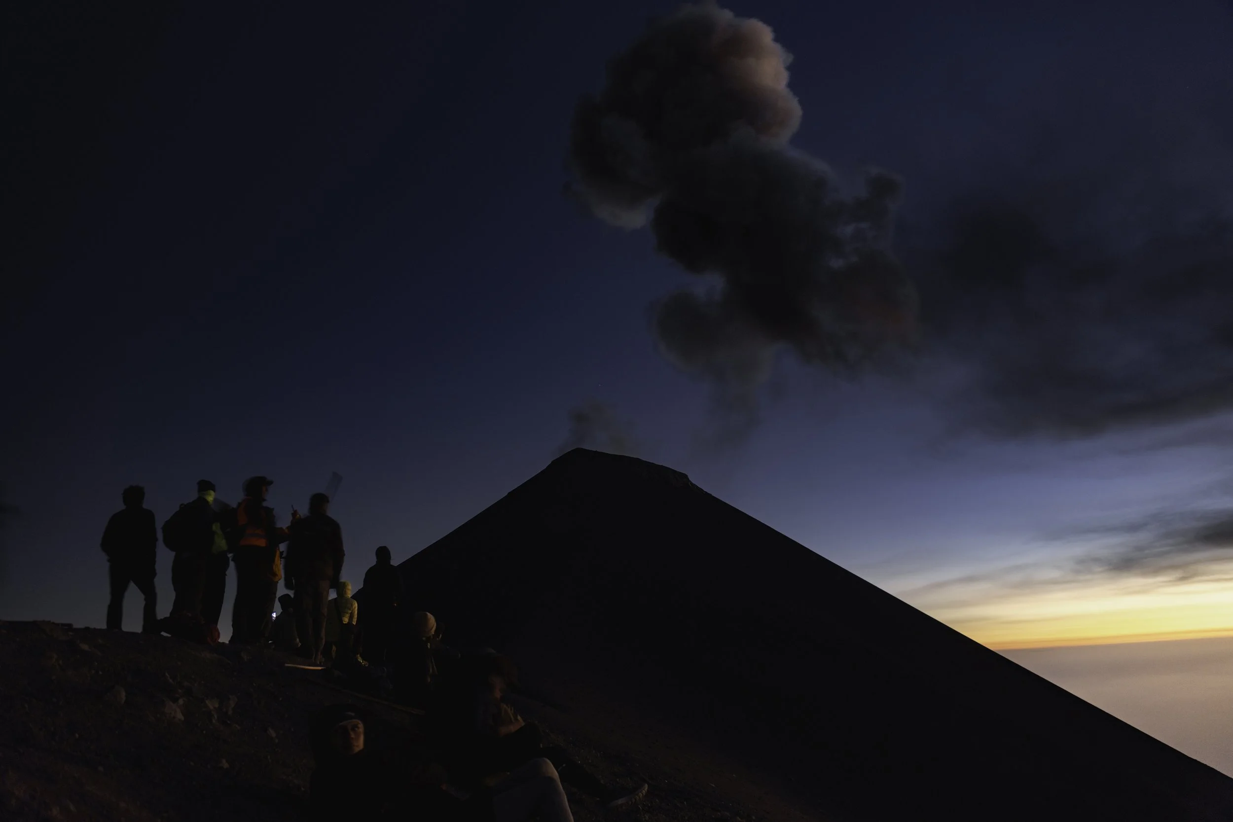 Group of people watching a volcanic eruption during dusk, with dark smoke billowing from the volcano's summit against a twilight sky.