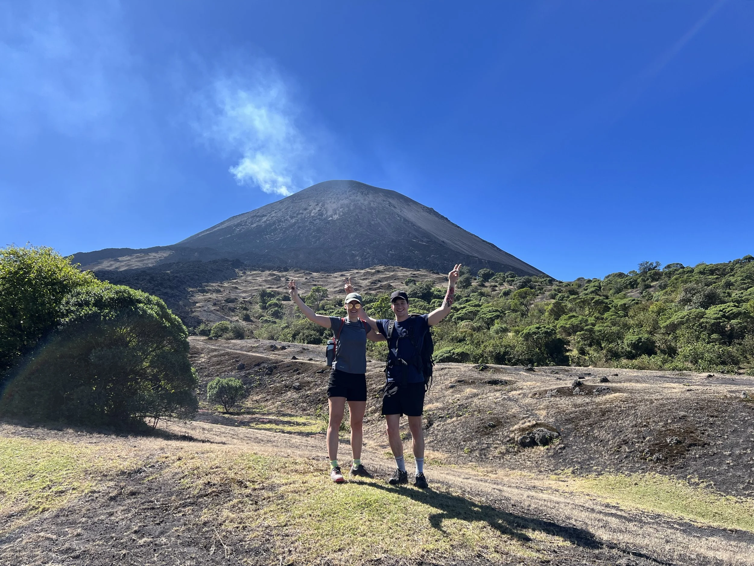 Two hikers standing in front of a volcanic mountain, smiling with arms raised, under a clear blue sky.