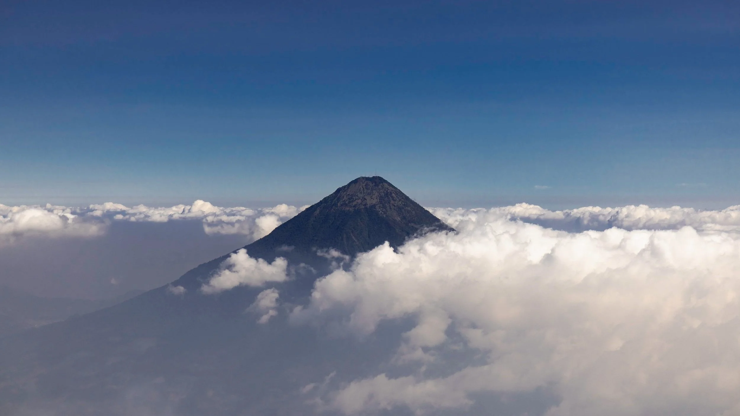 A tall volcano peak rising above a sea of clouds against a blue sky.