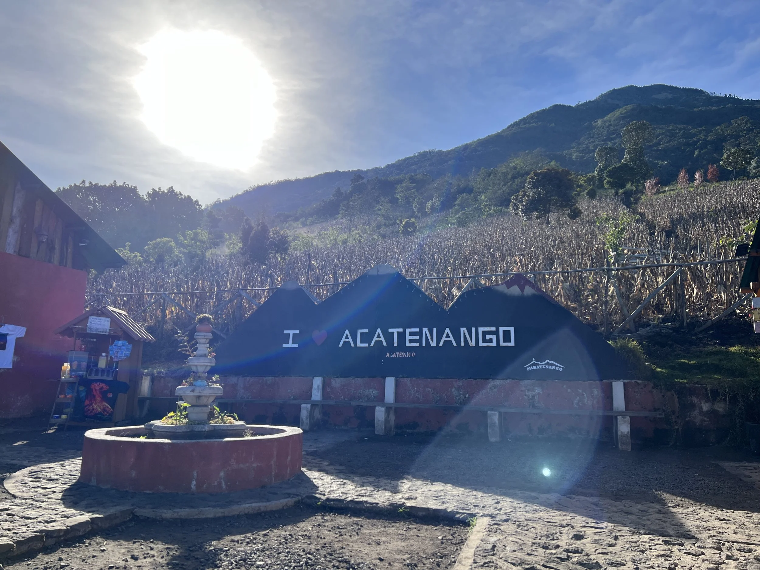 Sign that says 'I ❤️ ACATENANGO' in front of a hillside with dried crops and greenery, with mountains and a partly cloudy sky in the background.