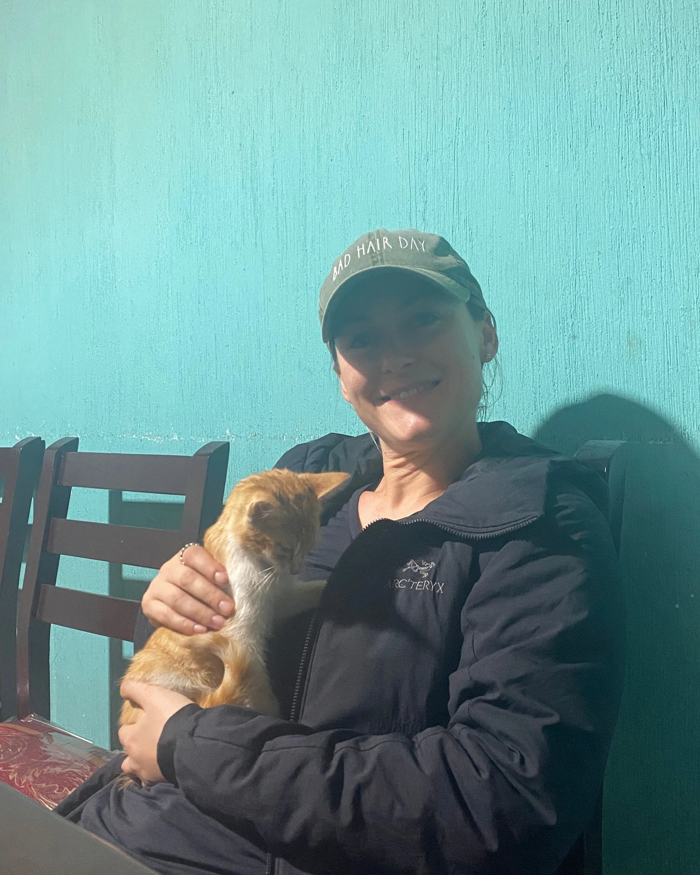 A woman sitting indoors on a chair, smiling, wearing a baseball cap that says 'BAD HAIR DAY' and a black Arc'teryx jacket. She is holding an orange and white cat close to her chest.
