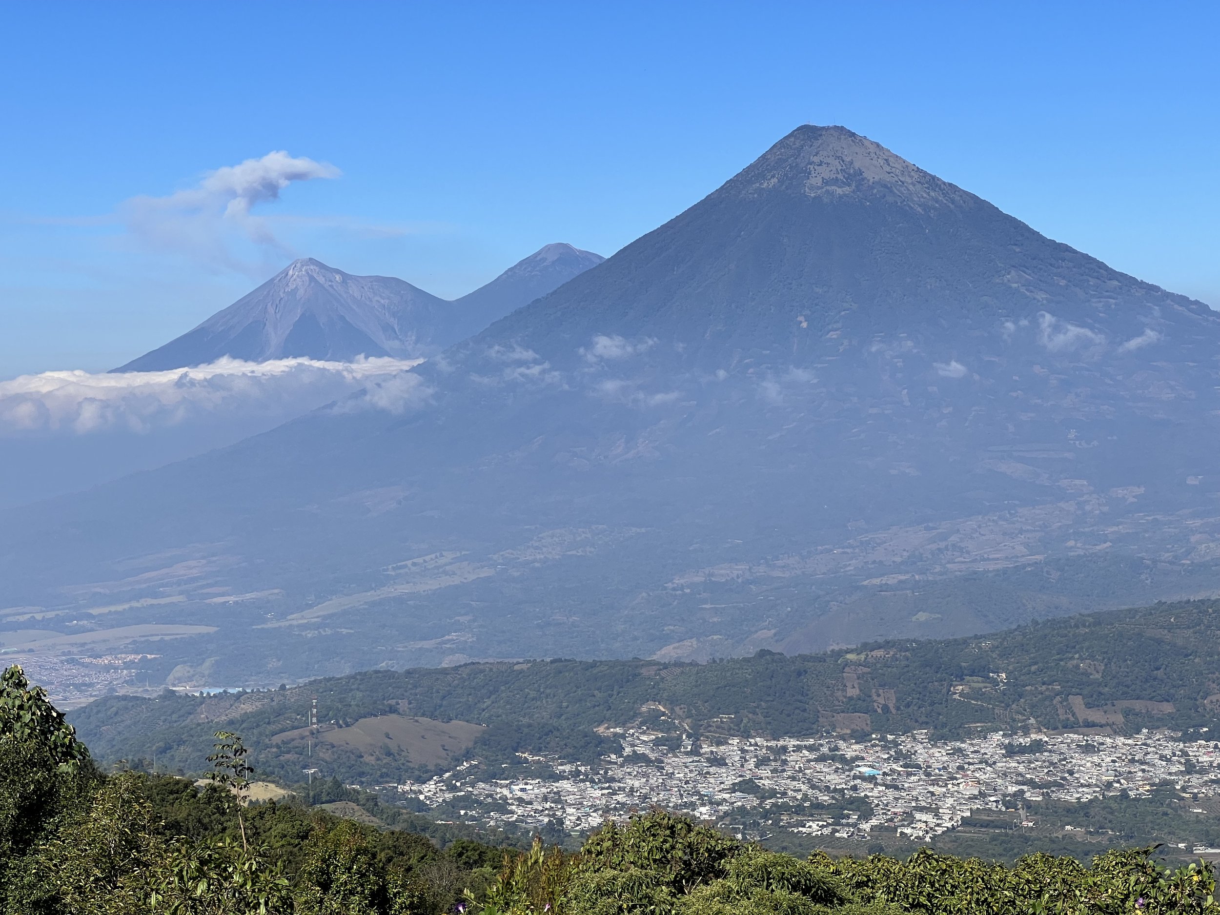 A scenic view of three volcanoes on a bright day, with a small town at the base and lush greenery in the foreground.
