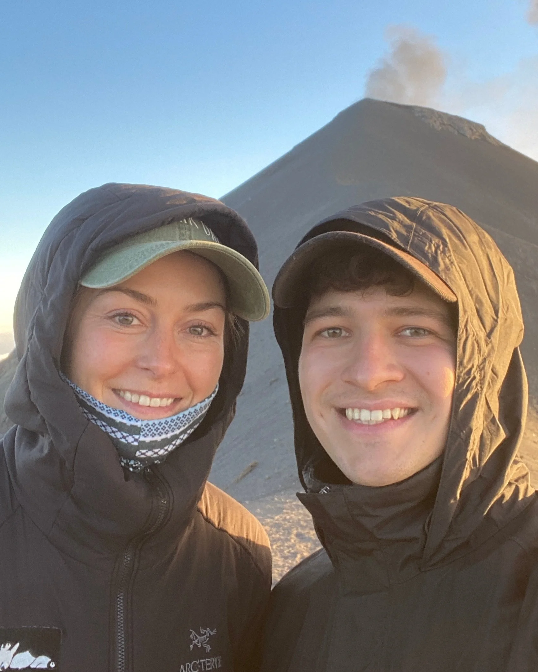 A smiling woman and man in outdoor jackets and hats are taking a selfie with a volcano erupting in the background.