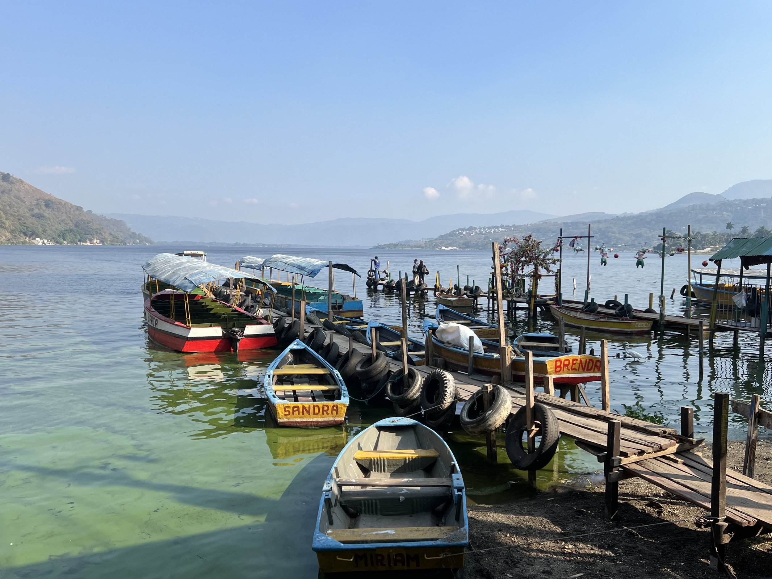 Boats docked along a wooden pier on a calm lake with mountains in the background under a clear blue sky.