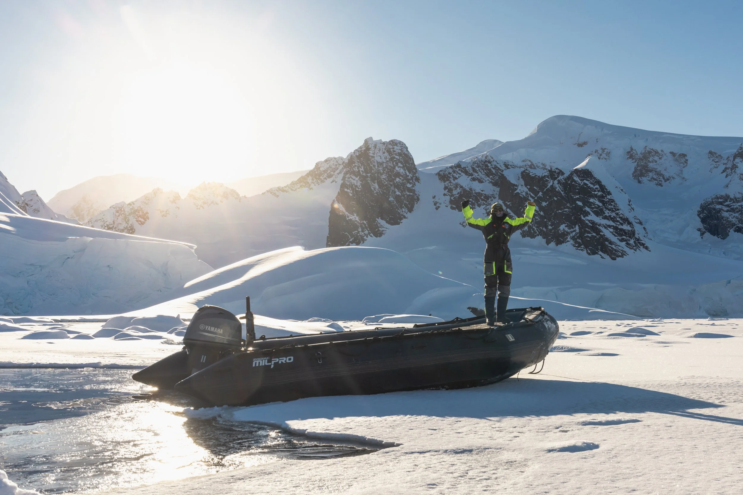 A person in winter gear standing on snow-covered ice in a mountainous landscape, with an inflatable boat and an outboard motor nearby, illuminated by bright sunlight.
