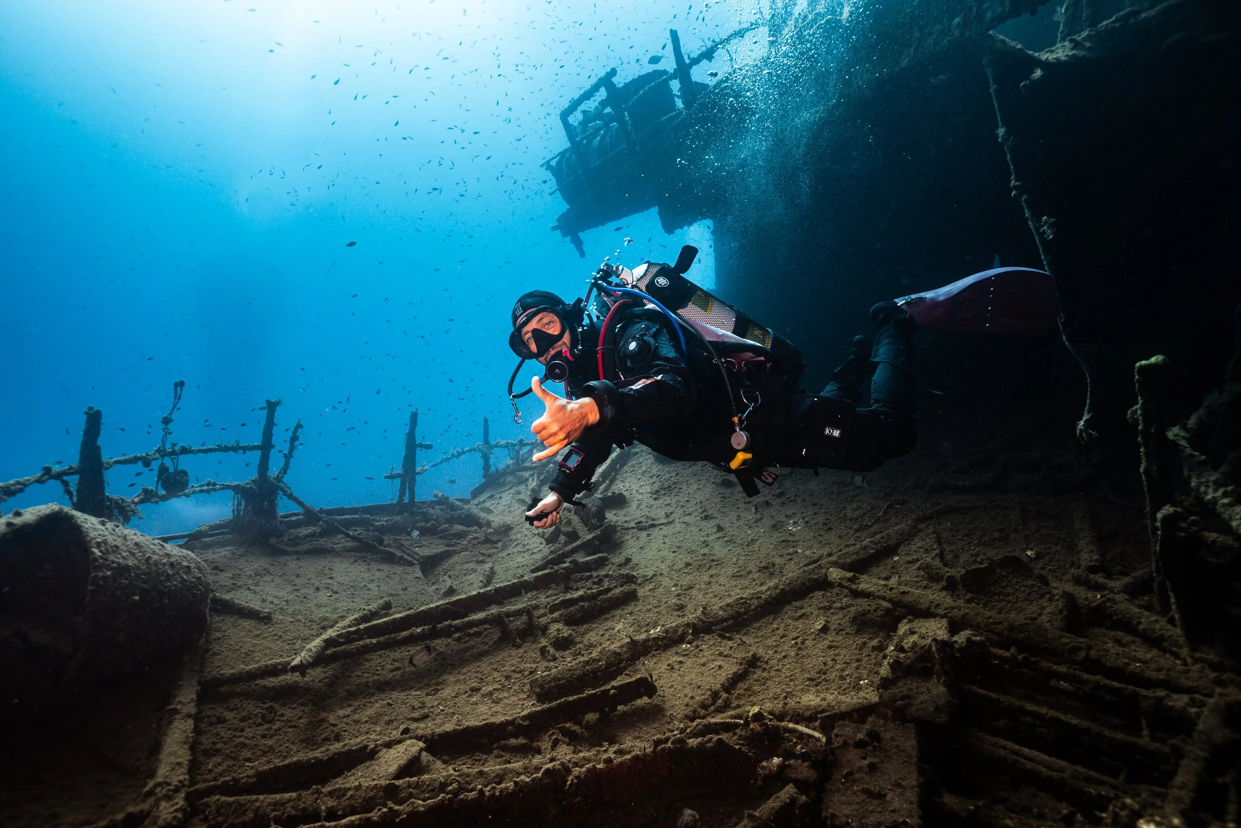 A scuba diver giving a thumbs-up underwater near a shipwreck, with the ocean and small fish in the background.