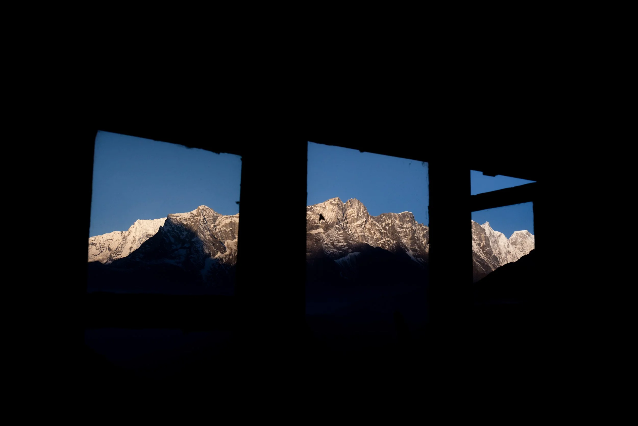 Snow-capped mountains seen through a window with a fabric shade partially pulled down, during daytime.