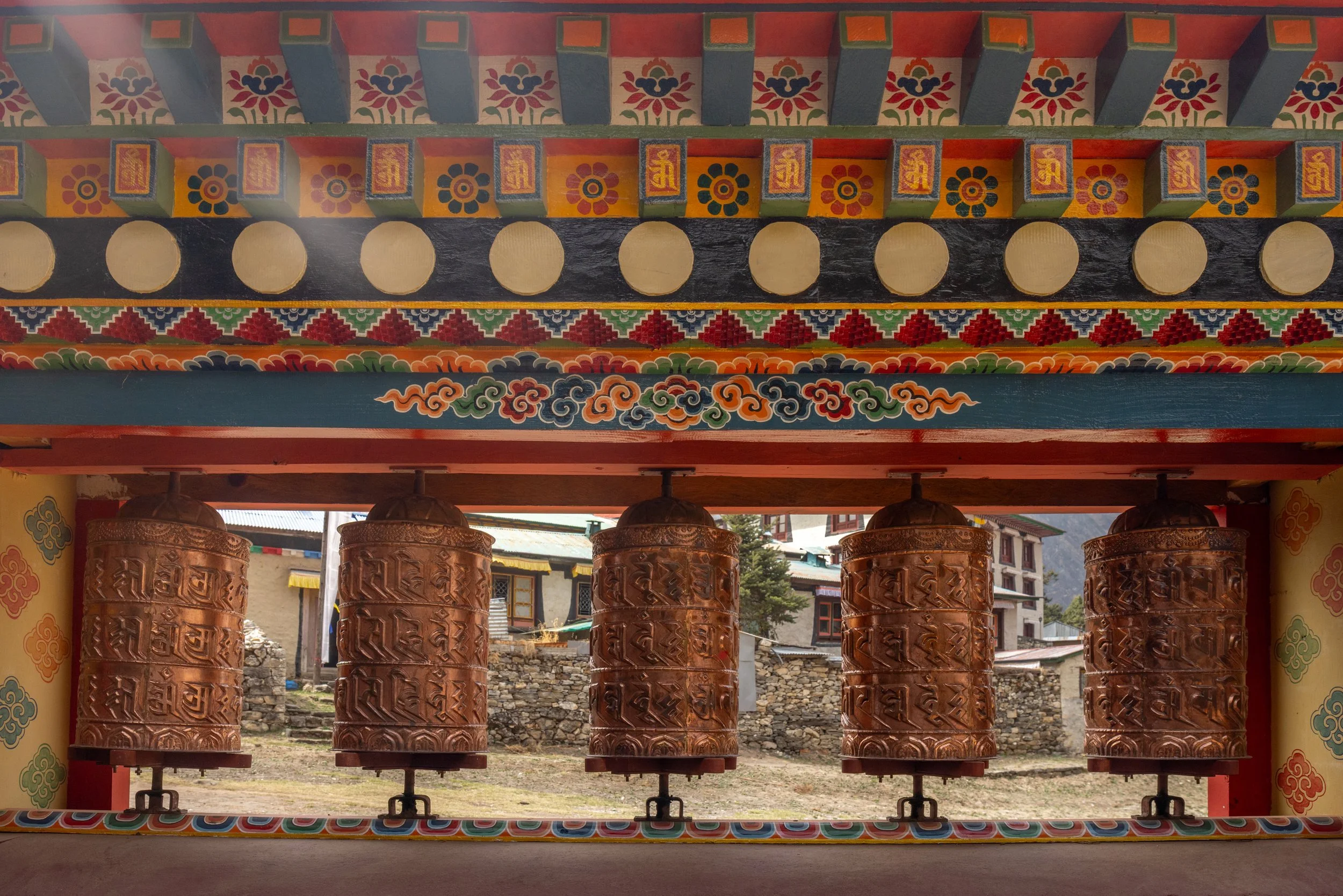 Four large Tibetan prayer wheels mounted on a colorful decorated structure, with a village and mountain background.