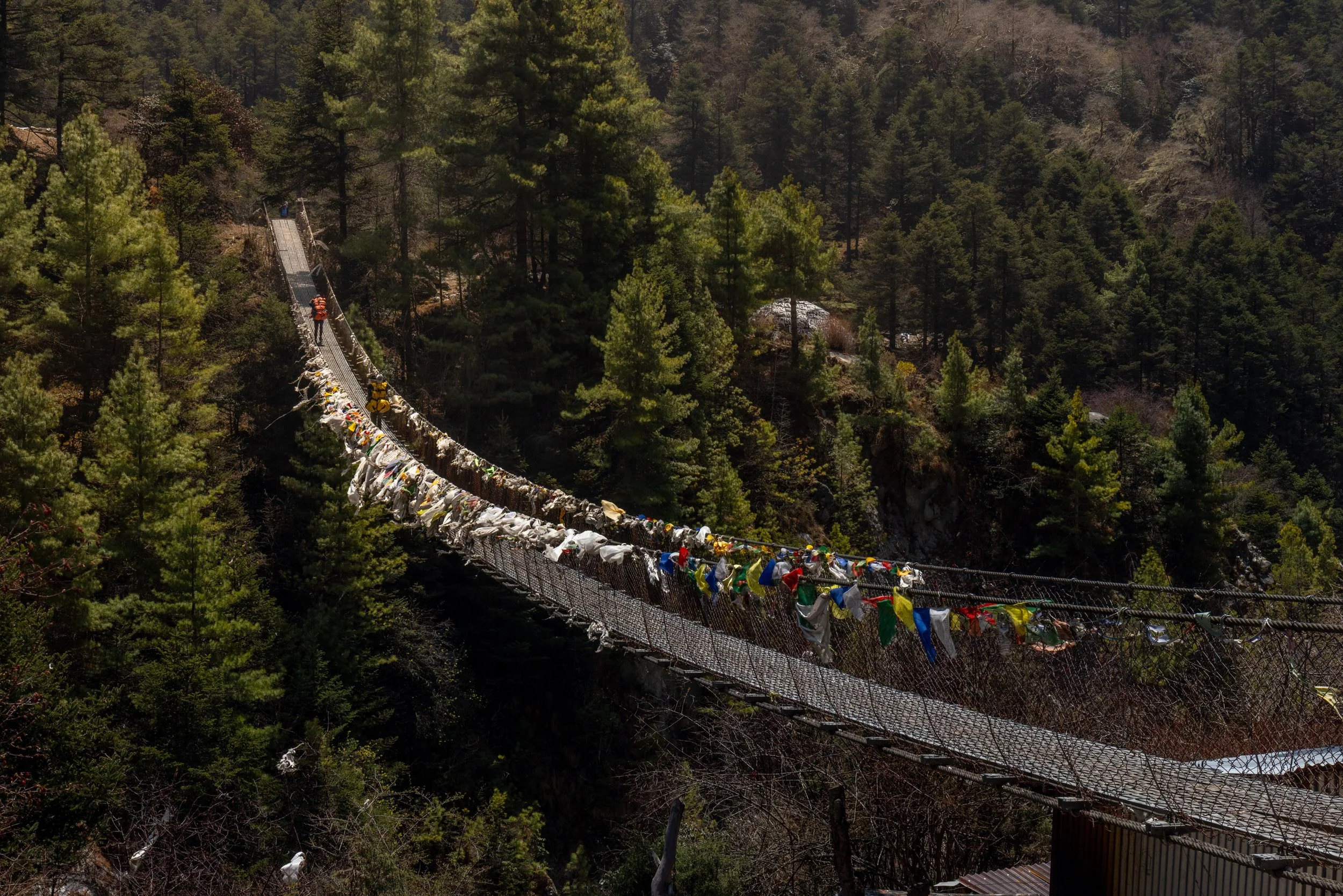 Suspension bridge in a forested area with trees, decorated with colorful cloth and flags, stretching across a valley.