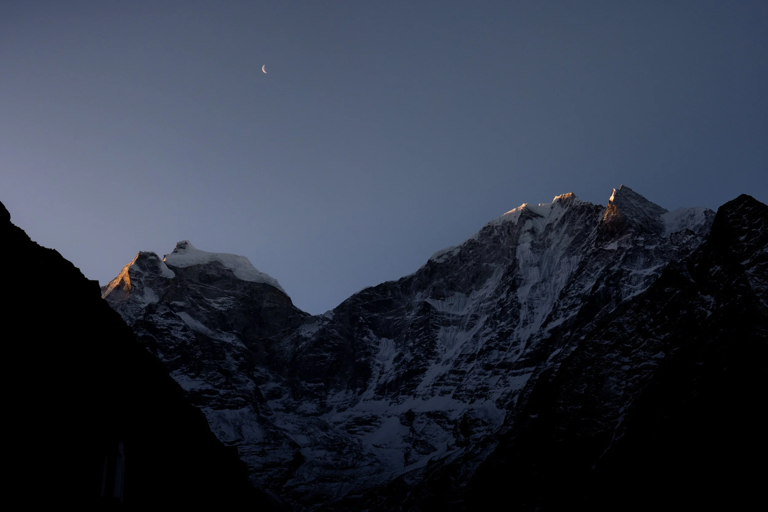 Nighttime mountain landscape with snow-capped peaks, crescent moon in the sky, and a clear dark blue sky.
