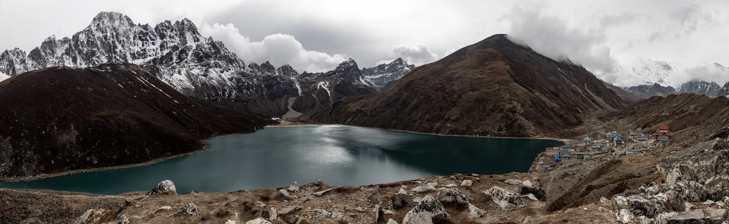 Mountain lake surrounded by rugged, rocky terrain with snow-capped peaks in the background and a small village on the right side.