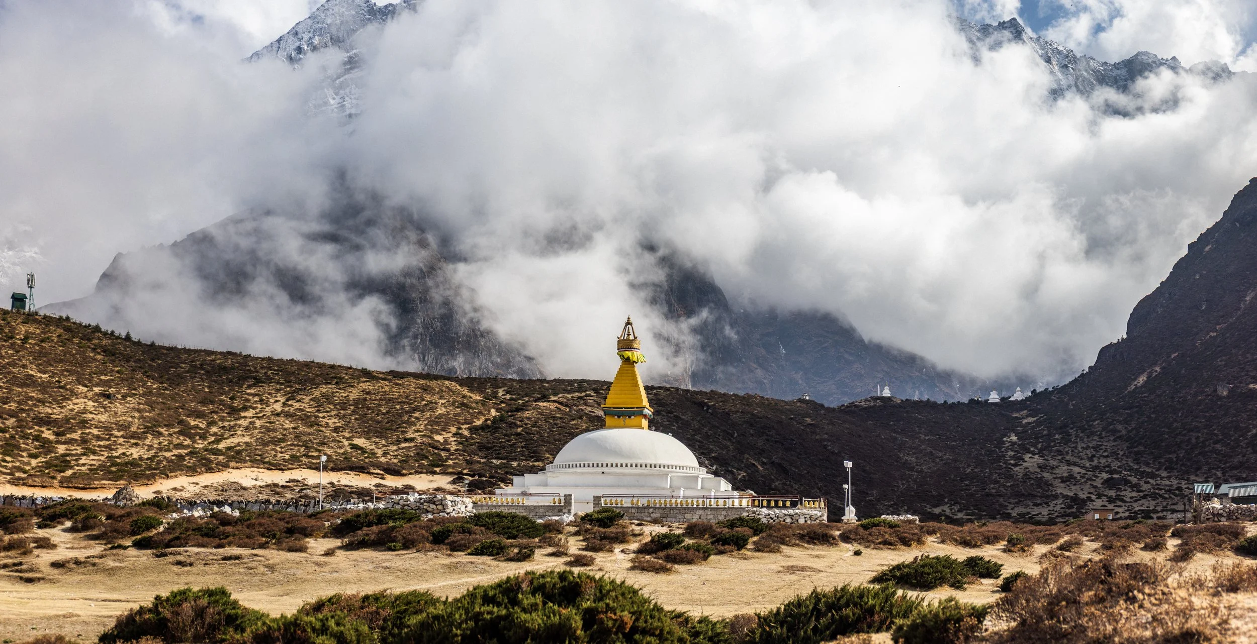 A white stupa with a golden spire in a mountainous landscape, surrounded by sparse vegetation and cloudy sky.