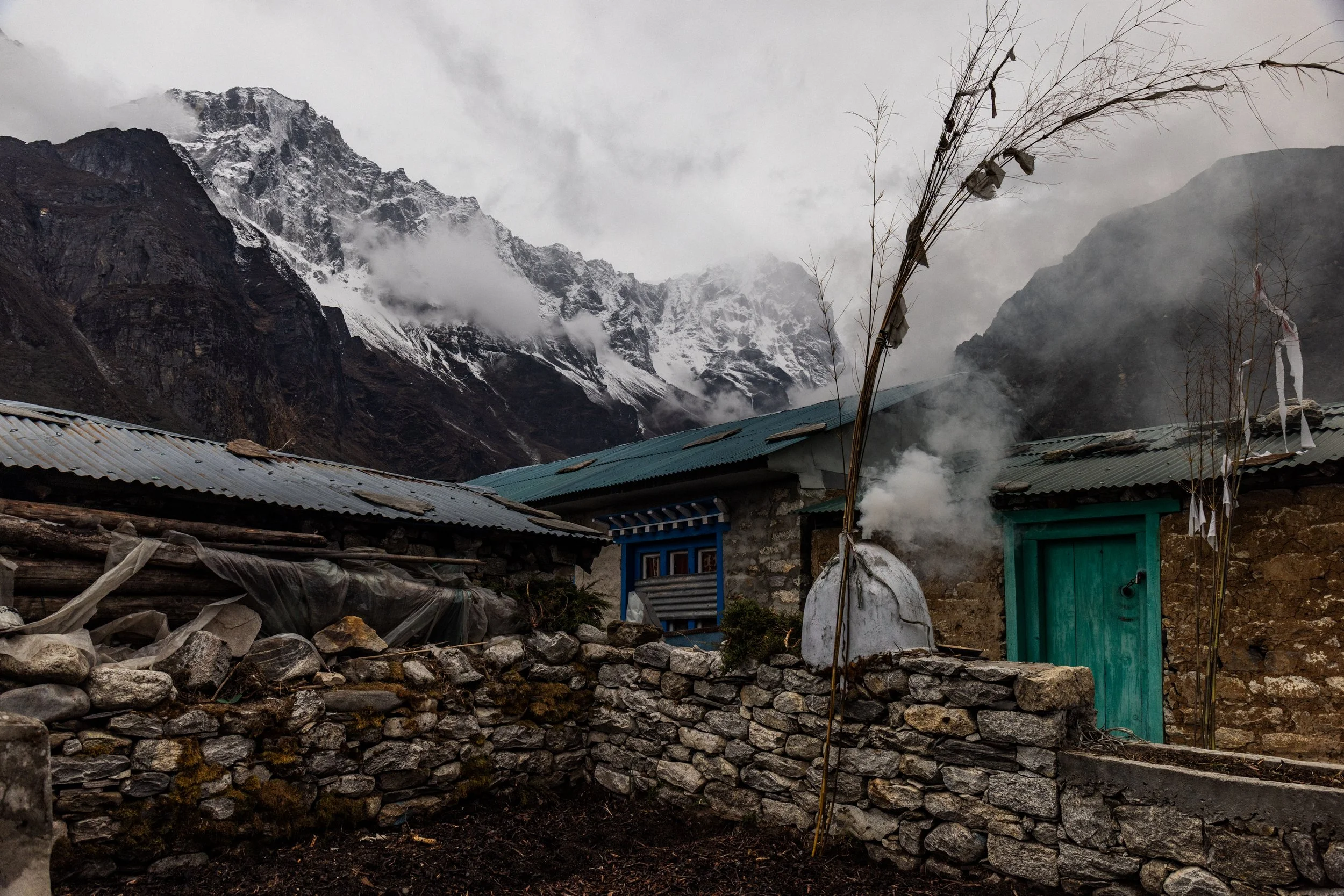 Mountain village with stone houses, some emitting steam, set against snow-capped peaks and cloudy sky.