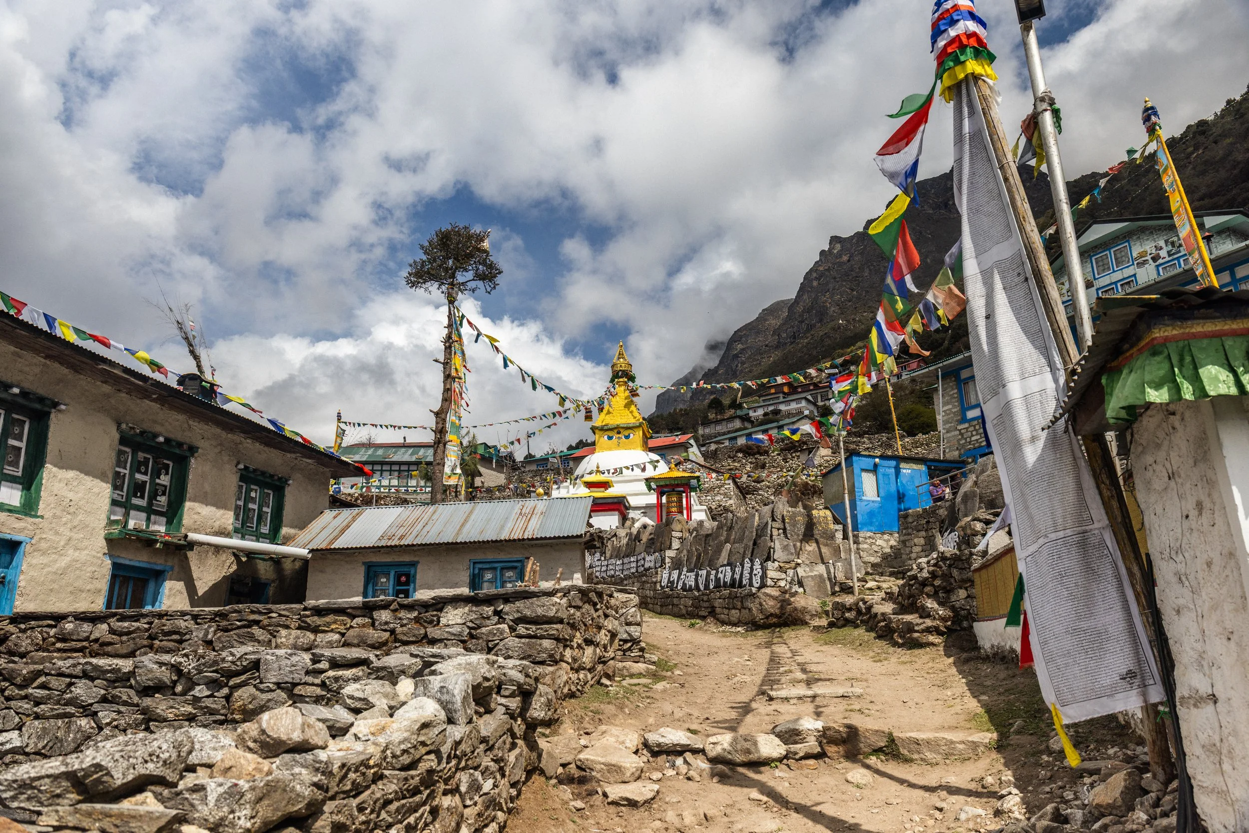 Himalayan village scene with colorful prayer flags, stone houses, and a Buddhist stupa at the center, set against a mountain backdrop with partly cloudy sky.