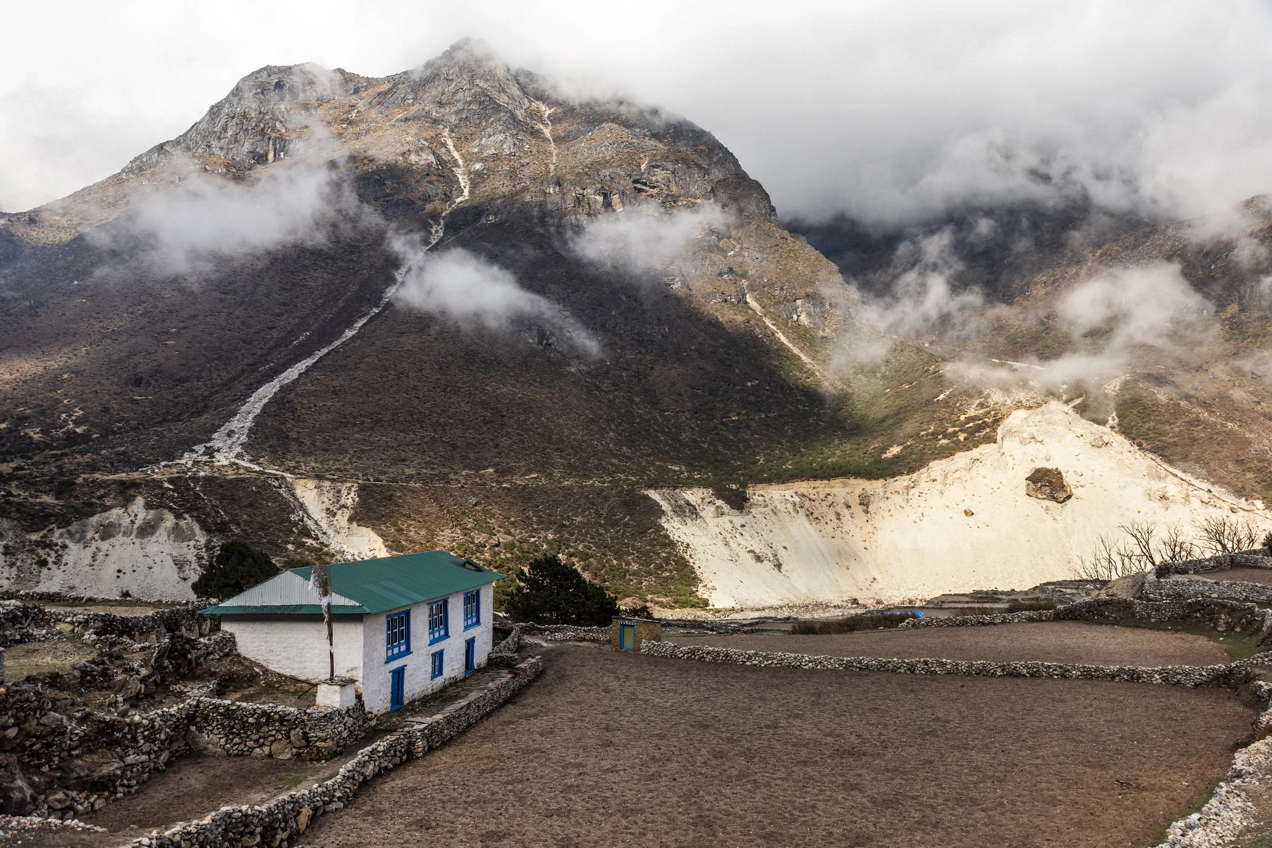 A small white house with a green roof and blue window frames in front of a mountainous landscape with cloudy skies.