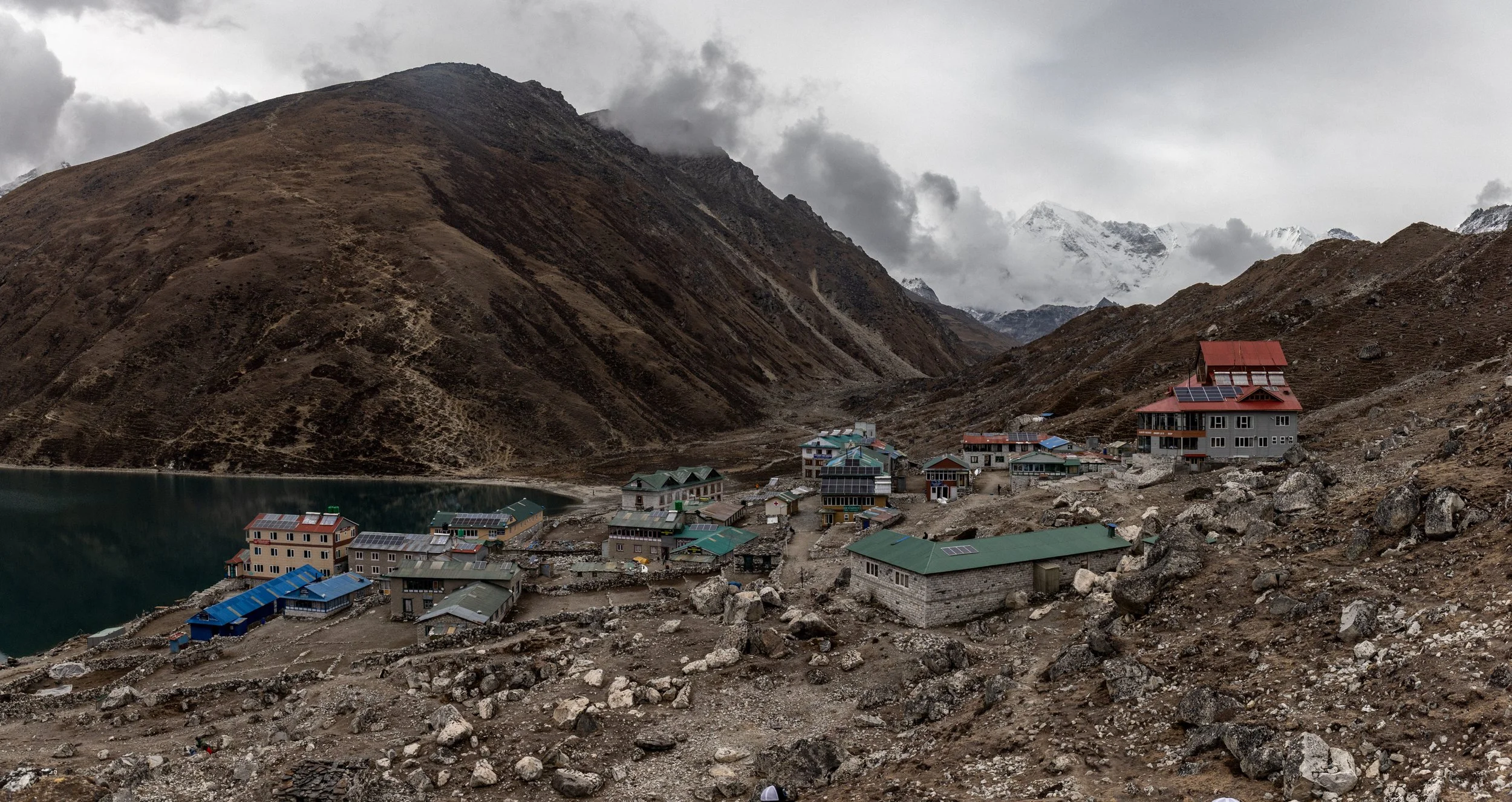 A mountain village with several buildings featuring green, blue, and red roofs near a lake, set against rugged mountain terrain with cloud-covered peaks in the background.