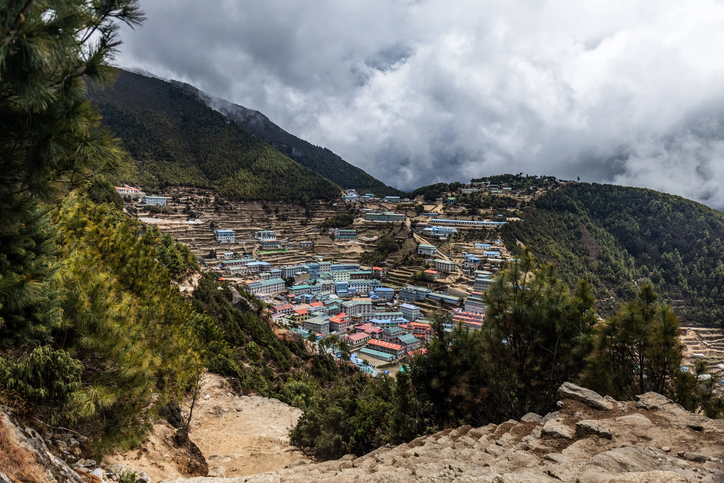 A mountain valley with numerous buildings, some with blue roofs, surrounded by lush green trees and terraced fields, with cloudy sky overhead.