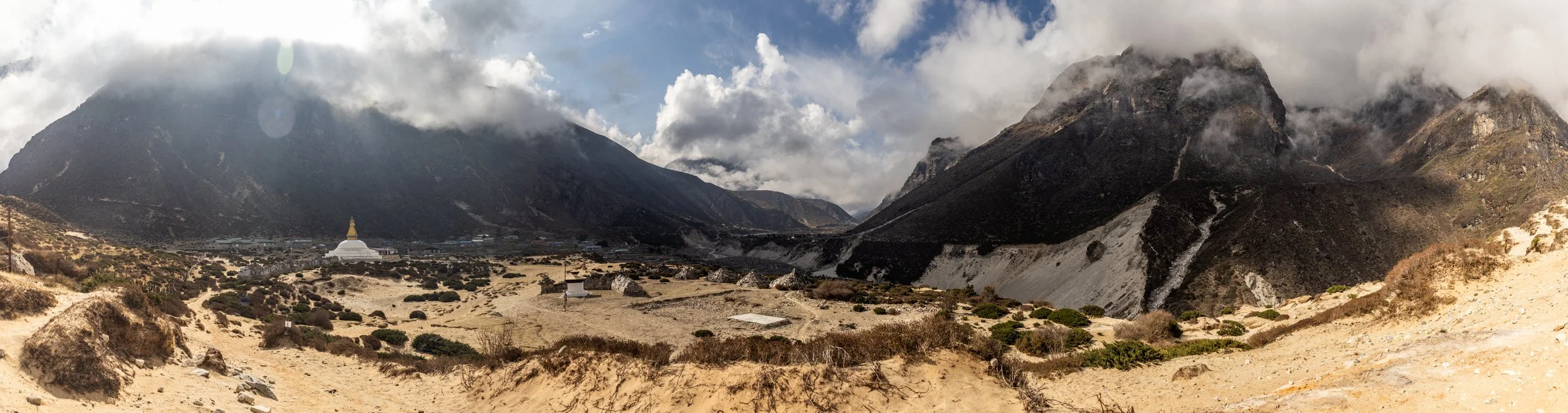 A panoramic view of a mountainous landscape with sandy foreground, scattered bushes, and clouds partially covering the mountain peaks.