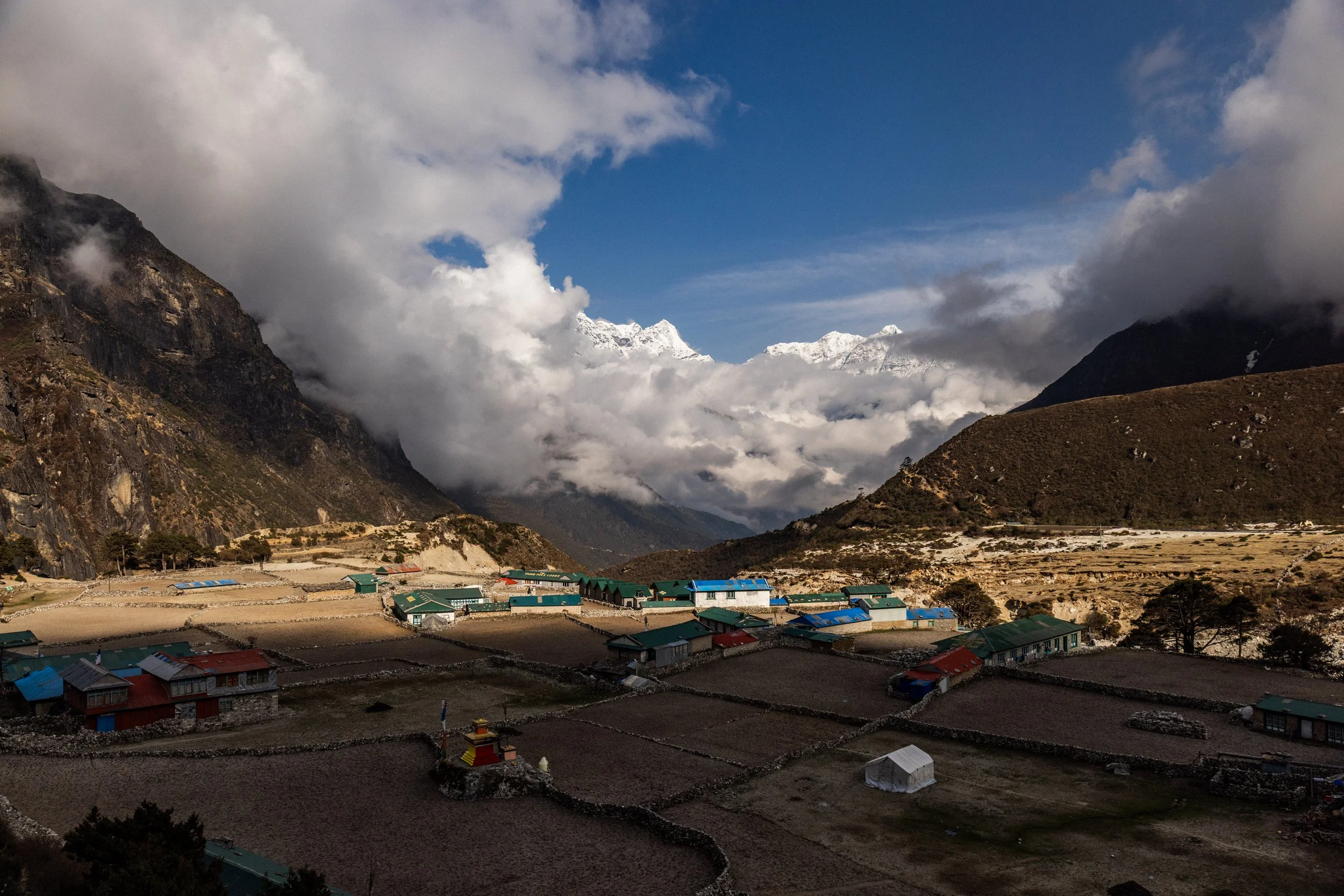 A village in a mountainous landscape with cloud-covered peaks in the background and small houses with colorful roofs in the foreground.