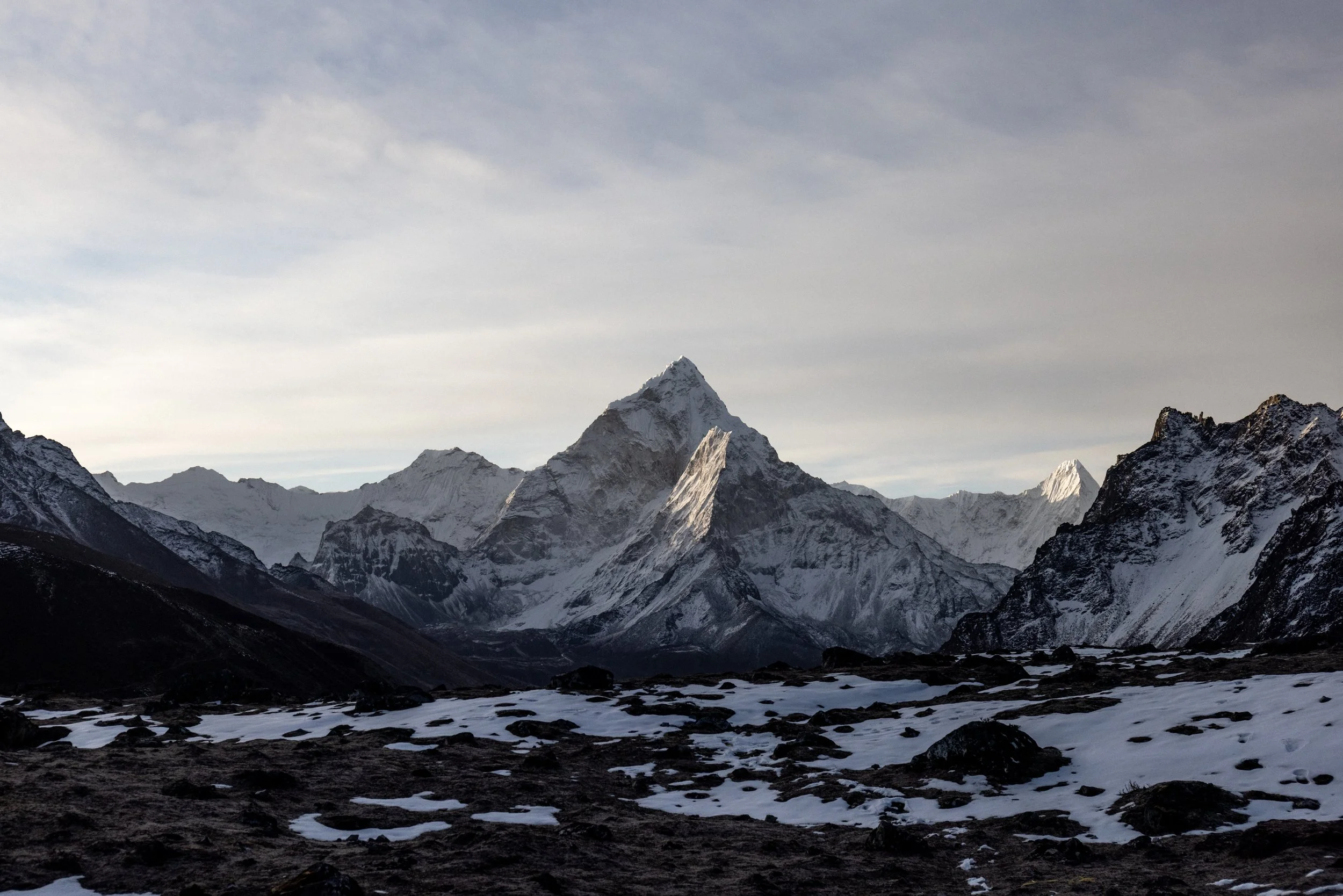 Snow-capped mountain range under a cloudy sky, with dark rocky terrain and patches of snow in the foreground.