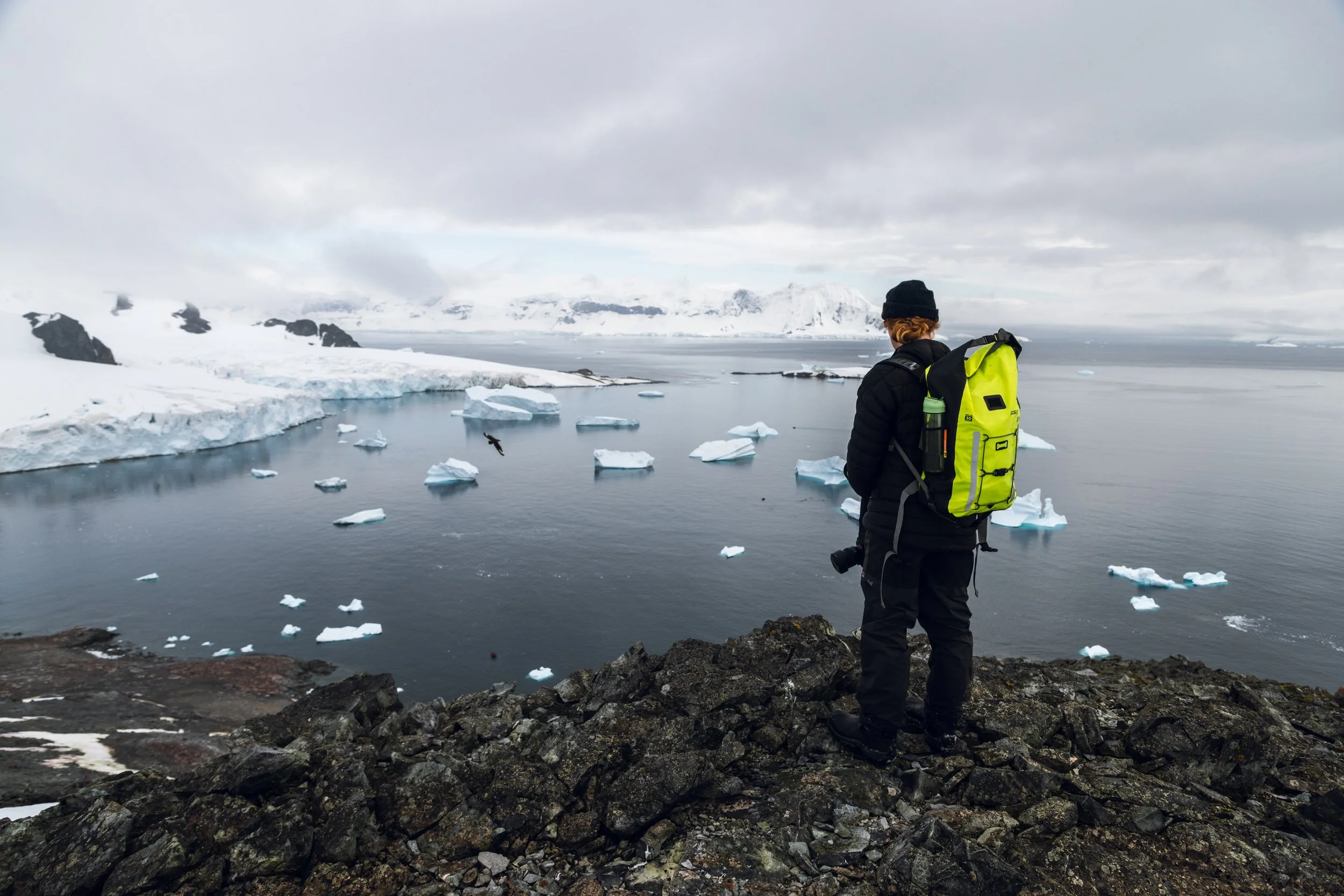 A person standing on rocky terrain overlooking a cold, icy landscape with floating icebergs in the water and snow-covered mountains in the background, under a cloudy sky.