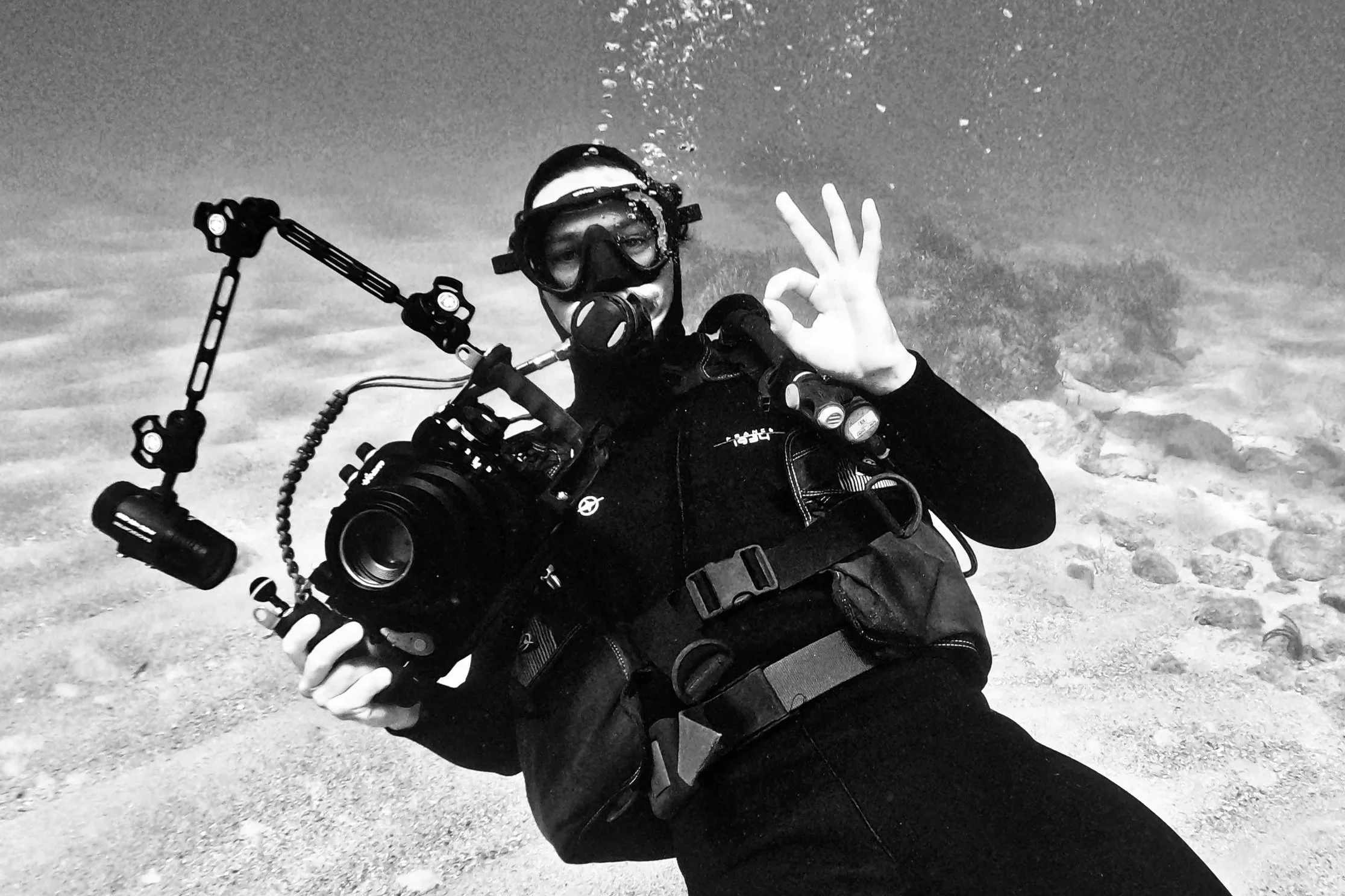 A diver in scuba gear making an OK hand gesture underwater, with a clear view of the ocean floor.