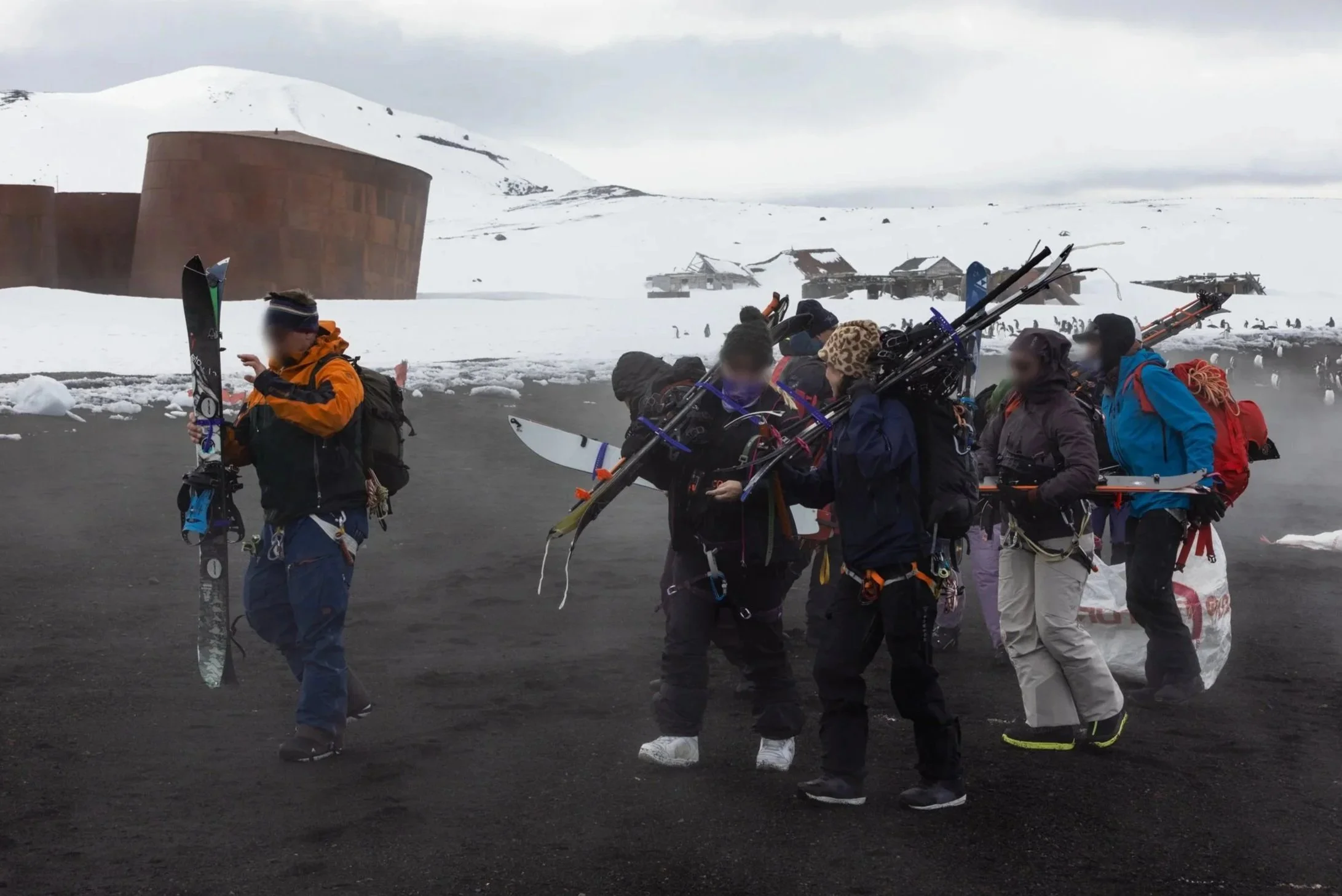 Group of people in winter gear carrying skis and snowboards on a snowy landscape, with abandoned buildings and snow-covered mountains in the background.