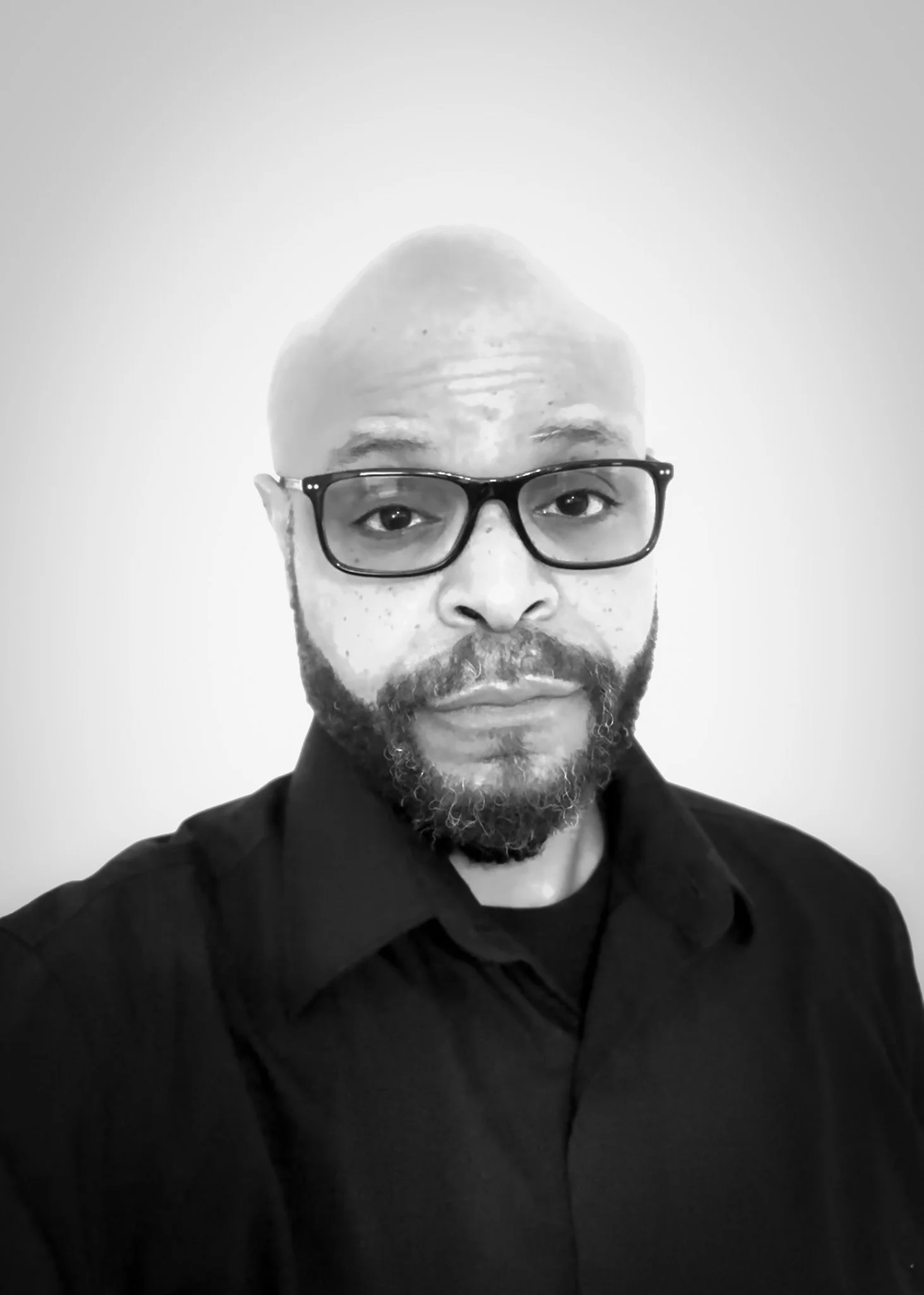 Black and white portrait of a man with glasses and a beard, wearing a dark shirt, looking directly at the camera.