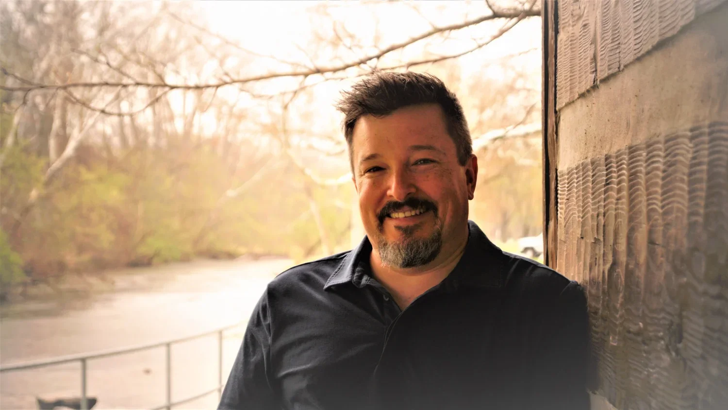 A man with dark hair and a beard smiling in front of a river with trees in the background during sunset.