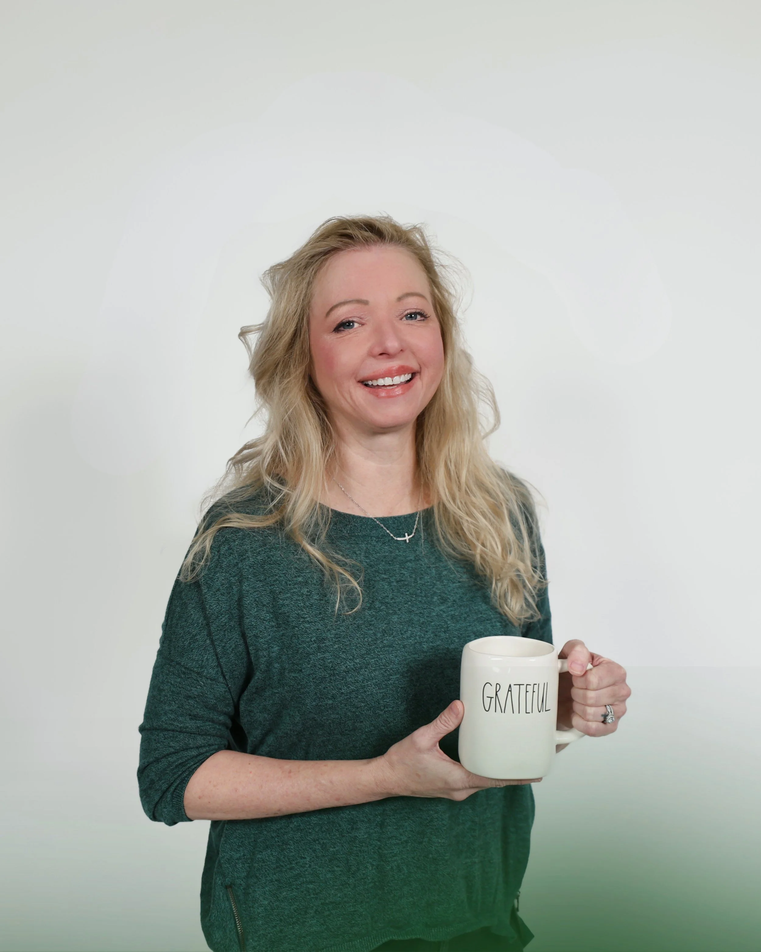 A blonde woman smiling while holding a white coffee mug with the word 'GRATEFUL' printed on it, standing in front of a plain light background.