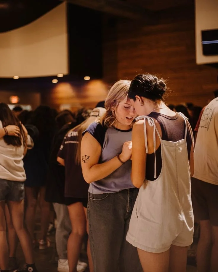 Two young women sharing an intimate moment, their foreheads touching, amidst a crowd at an indoor event or concert.