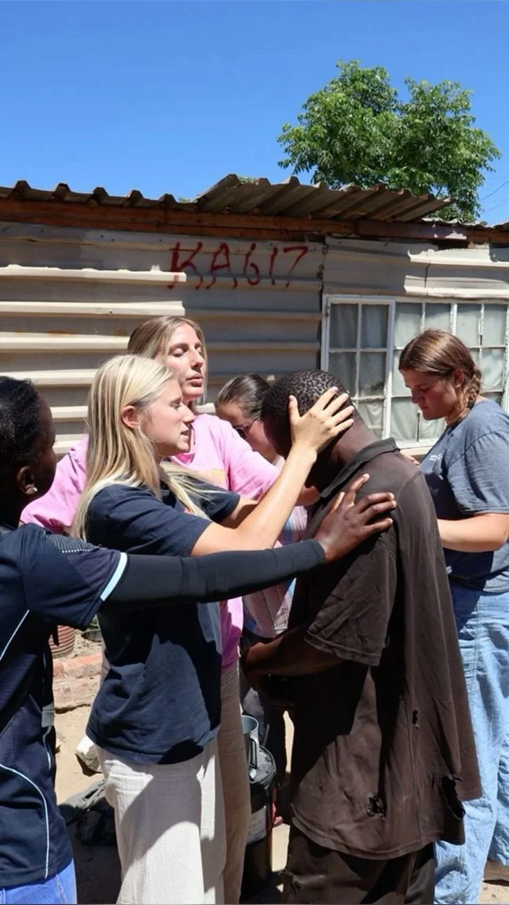 Group of women praying together outdoors in front of a metal building with graffiti and a tree, under a clear blue sky.