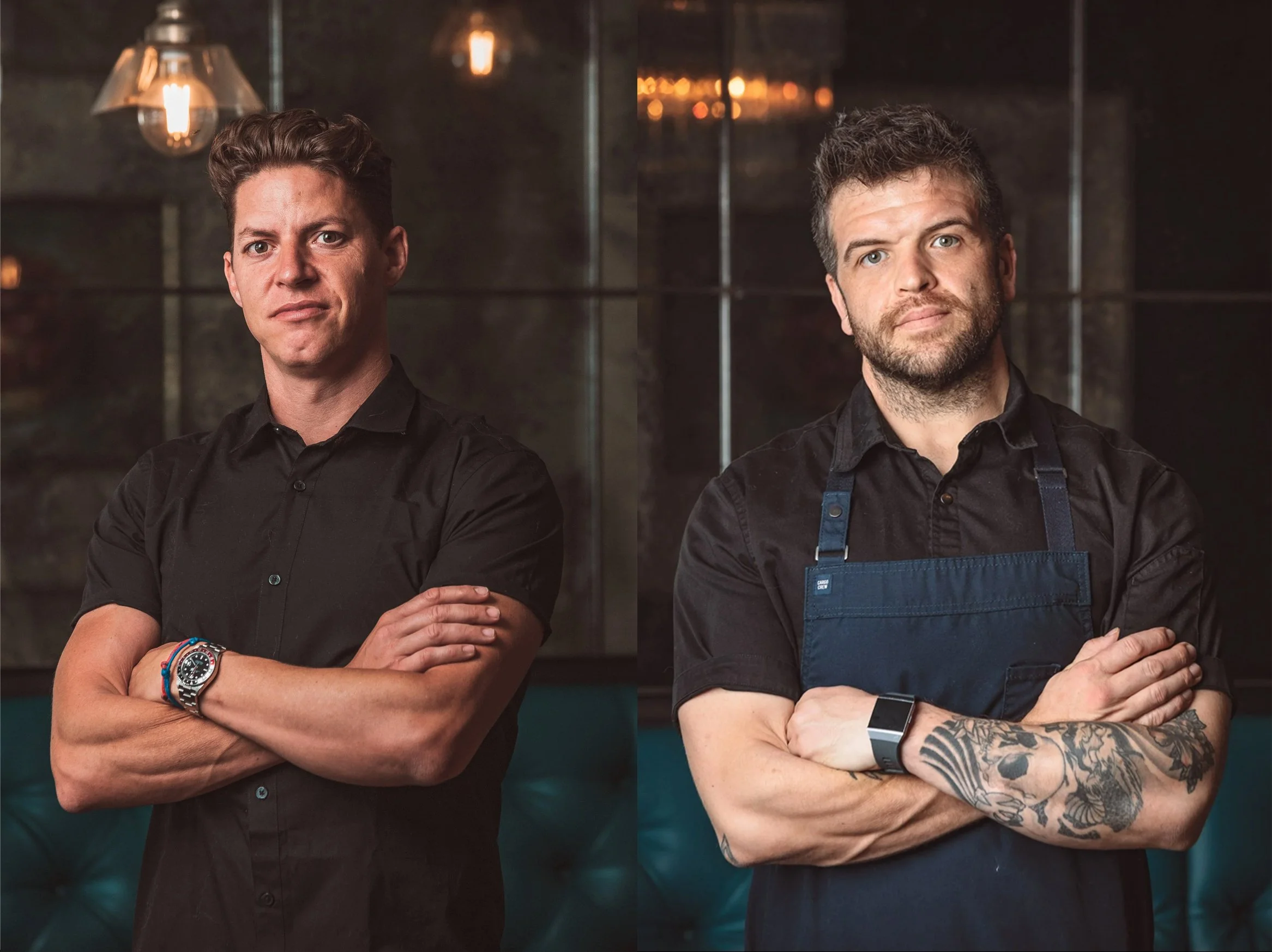 Two men with crossed arms pose for a photo, standing in front of a dark background with warm lighting. The man on the left wears a black shirt and a watch, while the man on the right wears a black shirt and an apron with tattoos on his arms.