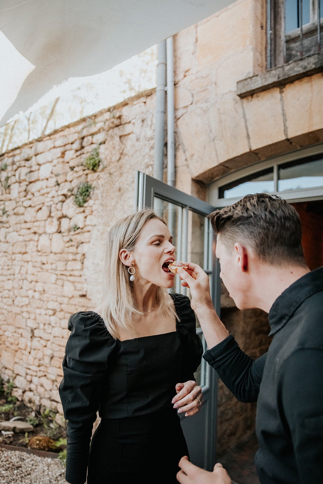 A woman with blonde hair and earrings is being fed a slice of pizza by a man with brown hair, standing in front of a brick wall with an open metal door.