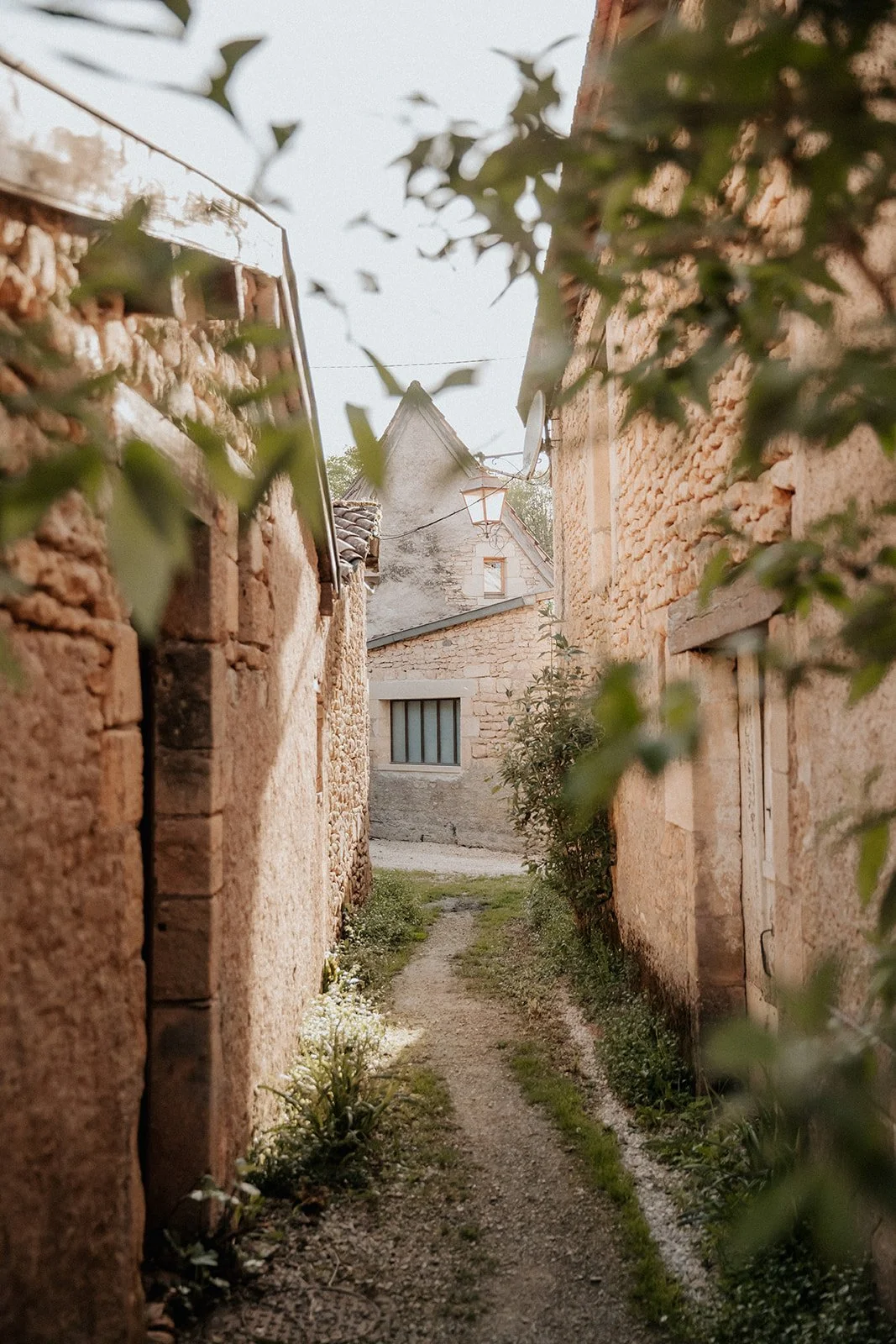 A narrow stone alleyway between old rustic buildings with ivy and greenery, leading to a small path and a house with a window, captured during daylight.