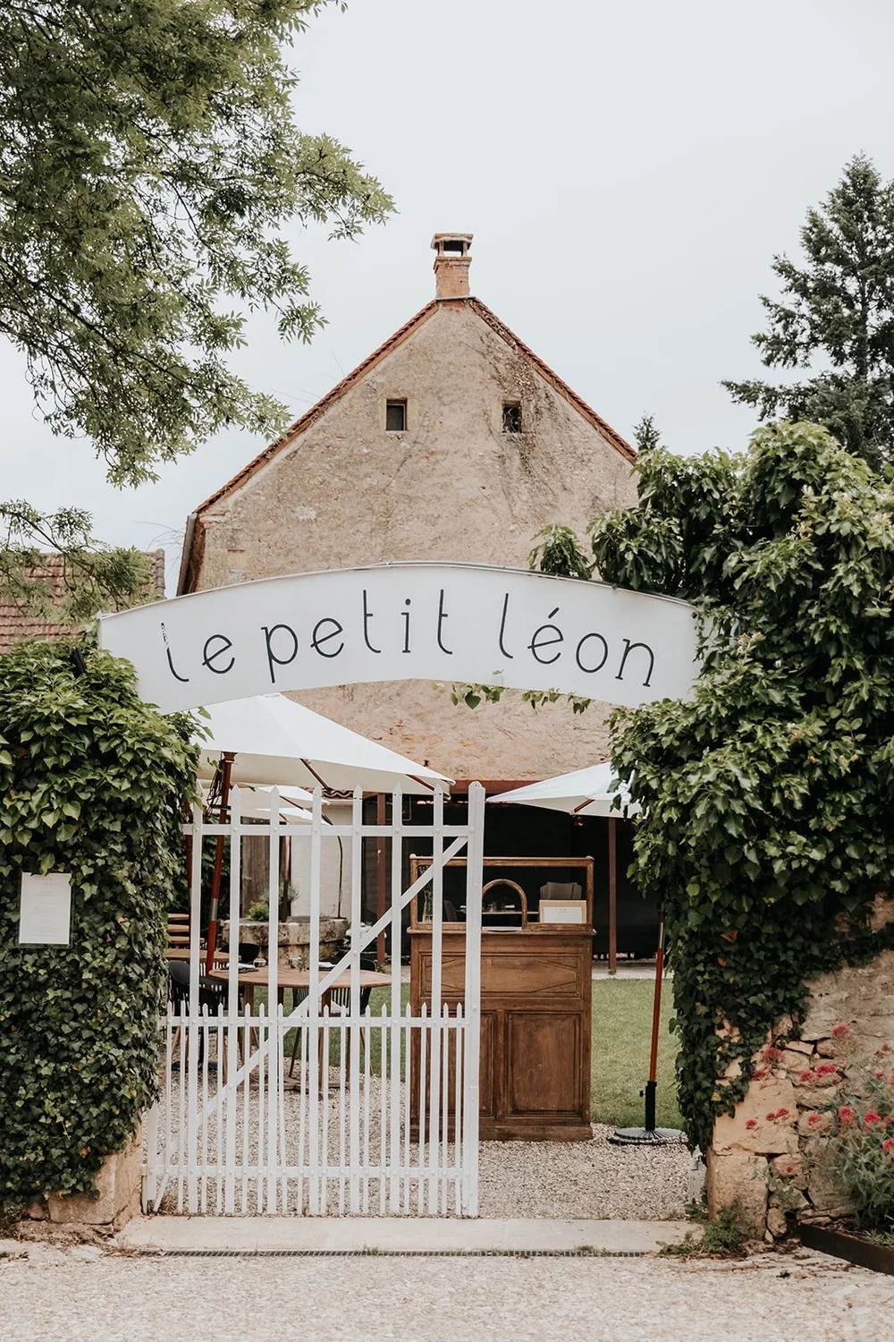Entrance gate with a sign that says 'le petit léon', leading to an outdoor seating area with umbrellas, trees, and an old stone building in the background.