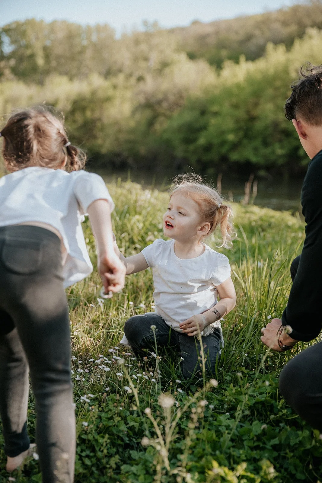 A young girl with red hair sitting on grass, holding hands with two children behind her, in a natural outdoor setting with greenery and trees.