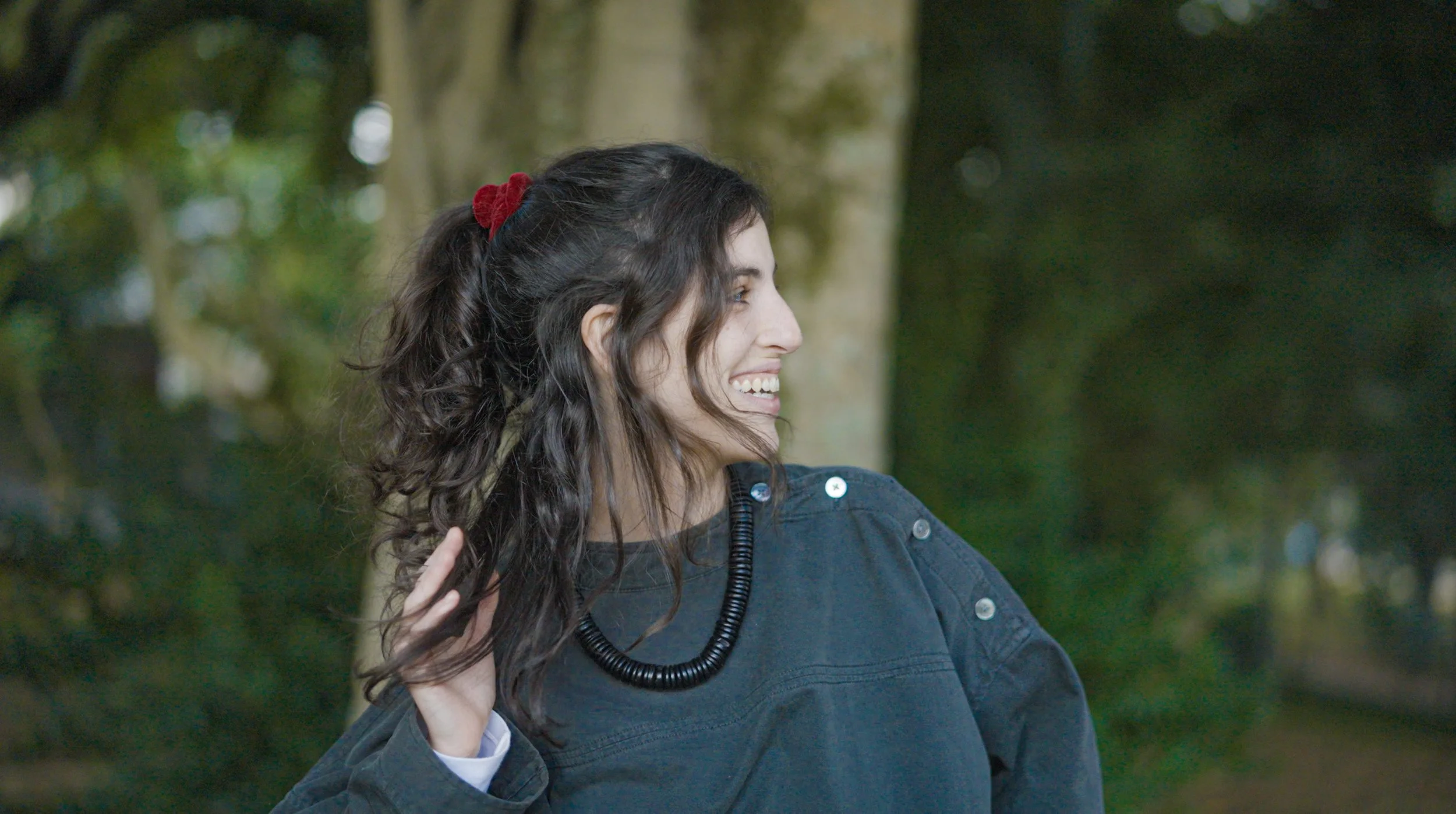 Foto de Julia Pasmanik, co-fundadora da Sementeira. A imagem revela mulher de perfil, sorridente, com cabelo escuro e cacheado, usando colar preto e casaco cinza, em um ambiente ao ar livre com árvores ao fundo.