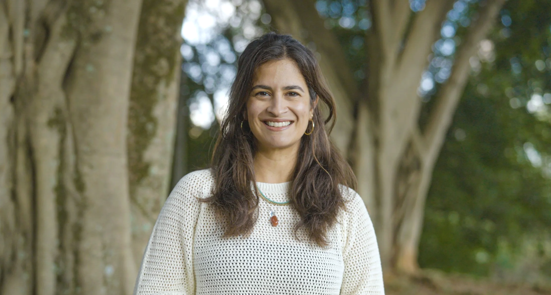Foto de Manuela Gambagorte, co-fundadora da Sementeira. A imagem revela uma mulher sorridente com cabelos castanhos ondulados, usando casaco branco de tricô, colar, em ambiente ao ar livre com árvores ao fundo.
