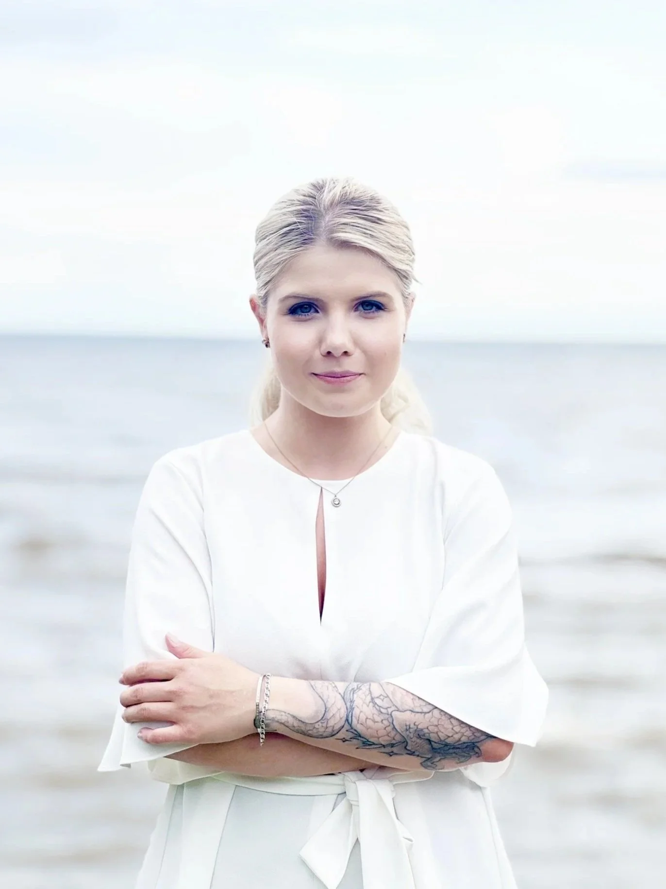 A woman with blonde hair, blue eyes, and tattoos on her left forearm, wearing a white blouse with a bow, stands with her arms crossed against a blurred background of a body of water and sky.