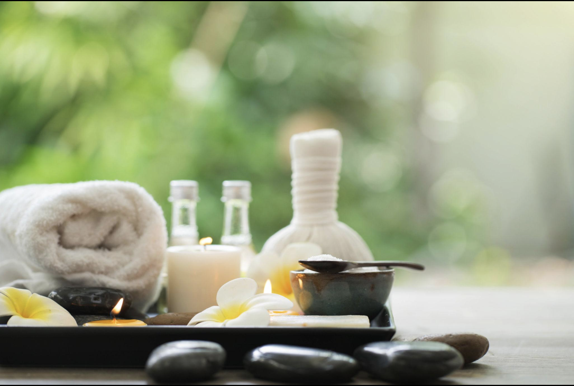 Spa setting with rolled towel, candles, small bottles, bowls, flower petals, and spa stones, with a green leafy background.