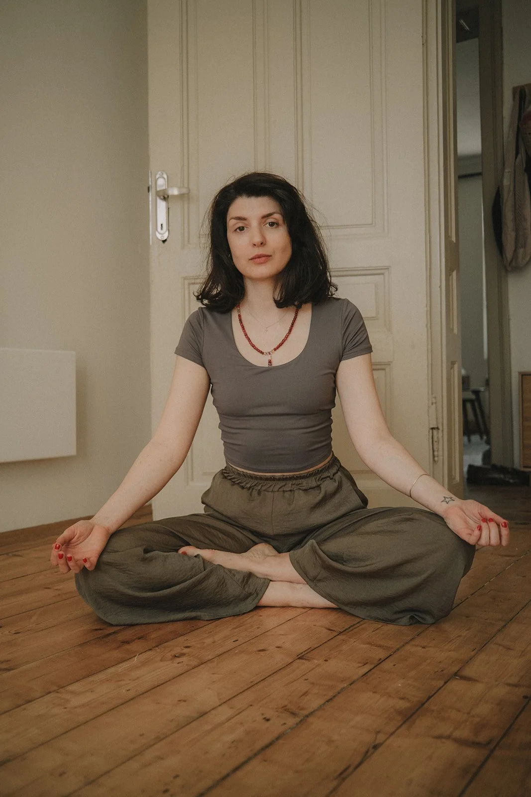 A woman with dark, shoulder-length hair and fair skin sits cross-legged on a wooden floor, meditating in front of a closed door in a home interior.