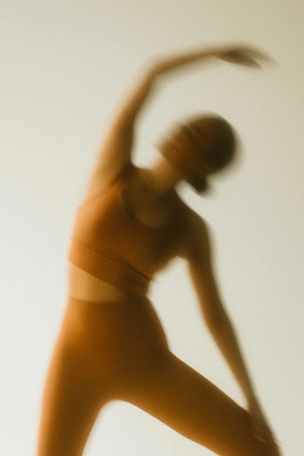 A blurred silhouette of a person in a dance pose with one arm raised and head tilted downward against a plain background.