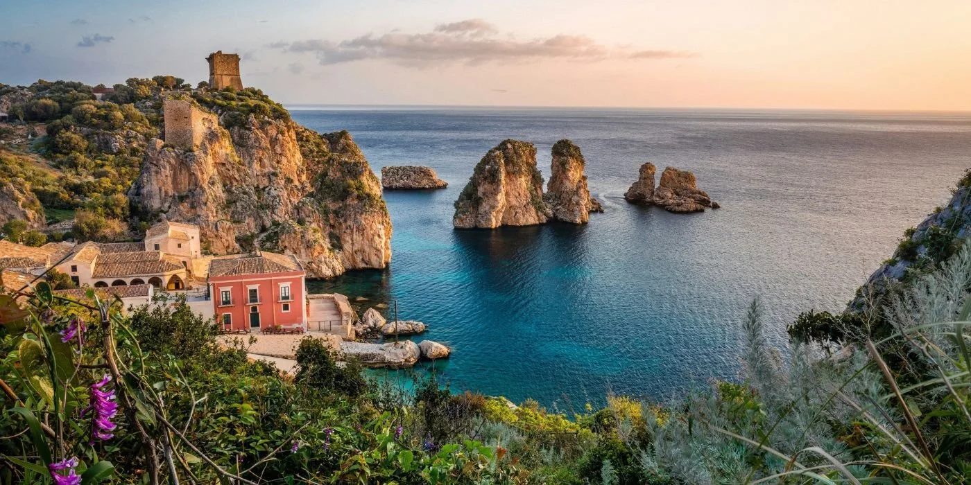Paesaggio costiero con scogliere rocciose, case colorate, vegetazione e il mare al tramonto.