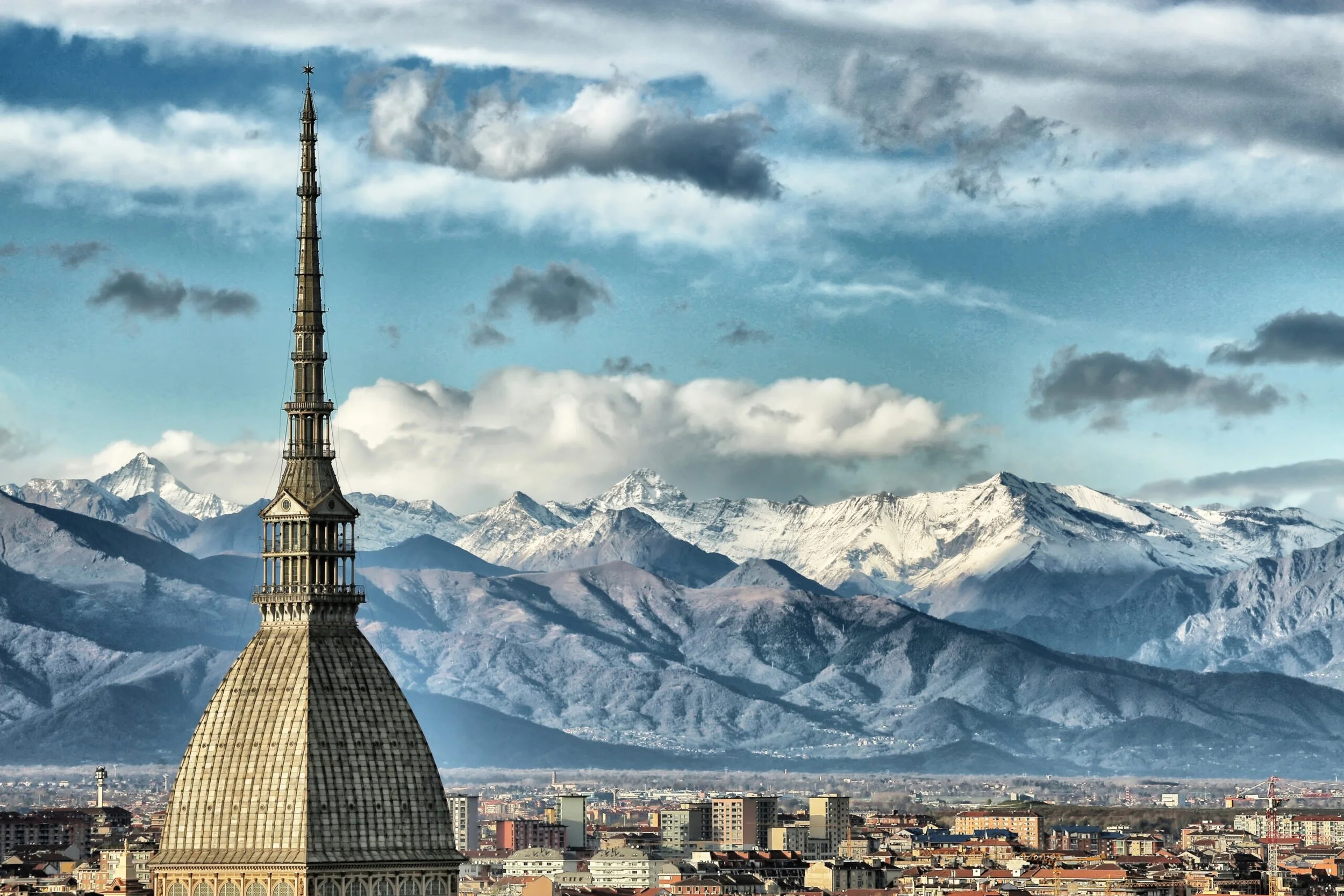 Una torre con tetto a ombrello in stile barocco in primo piano, con montagne innevate e un cielo con nuvole sullo sfondo